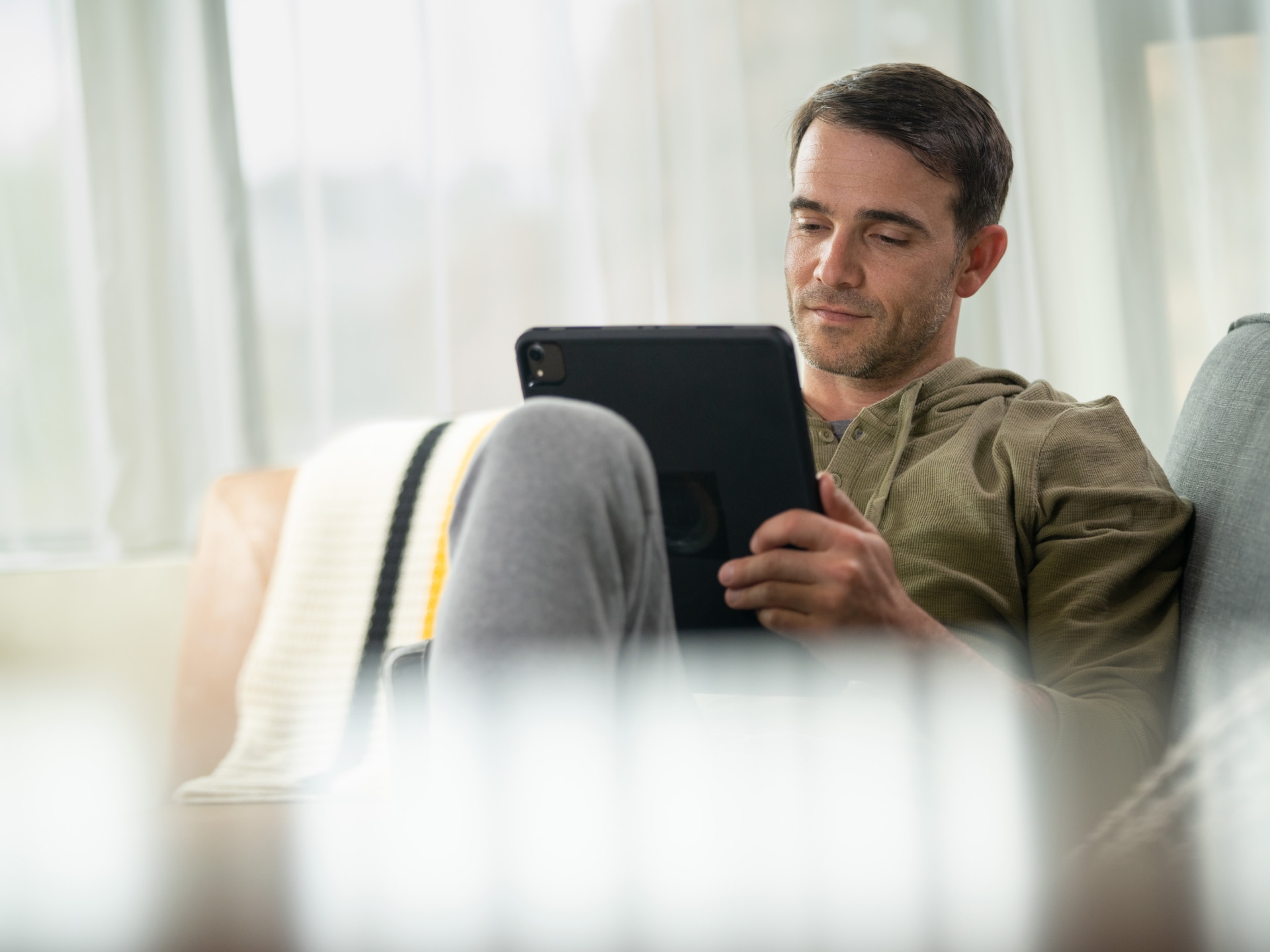 A Man Relaxing on a Sofa and Looking at a Tablet Screen with a Blanket on His Lap