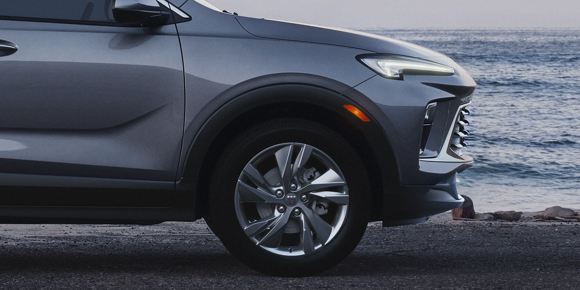  A Metallic Gray Buick Encore GX SUV Parked by the Ocean with Its Headlight on and the Buick Logo Visible on the Wheel