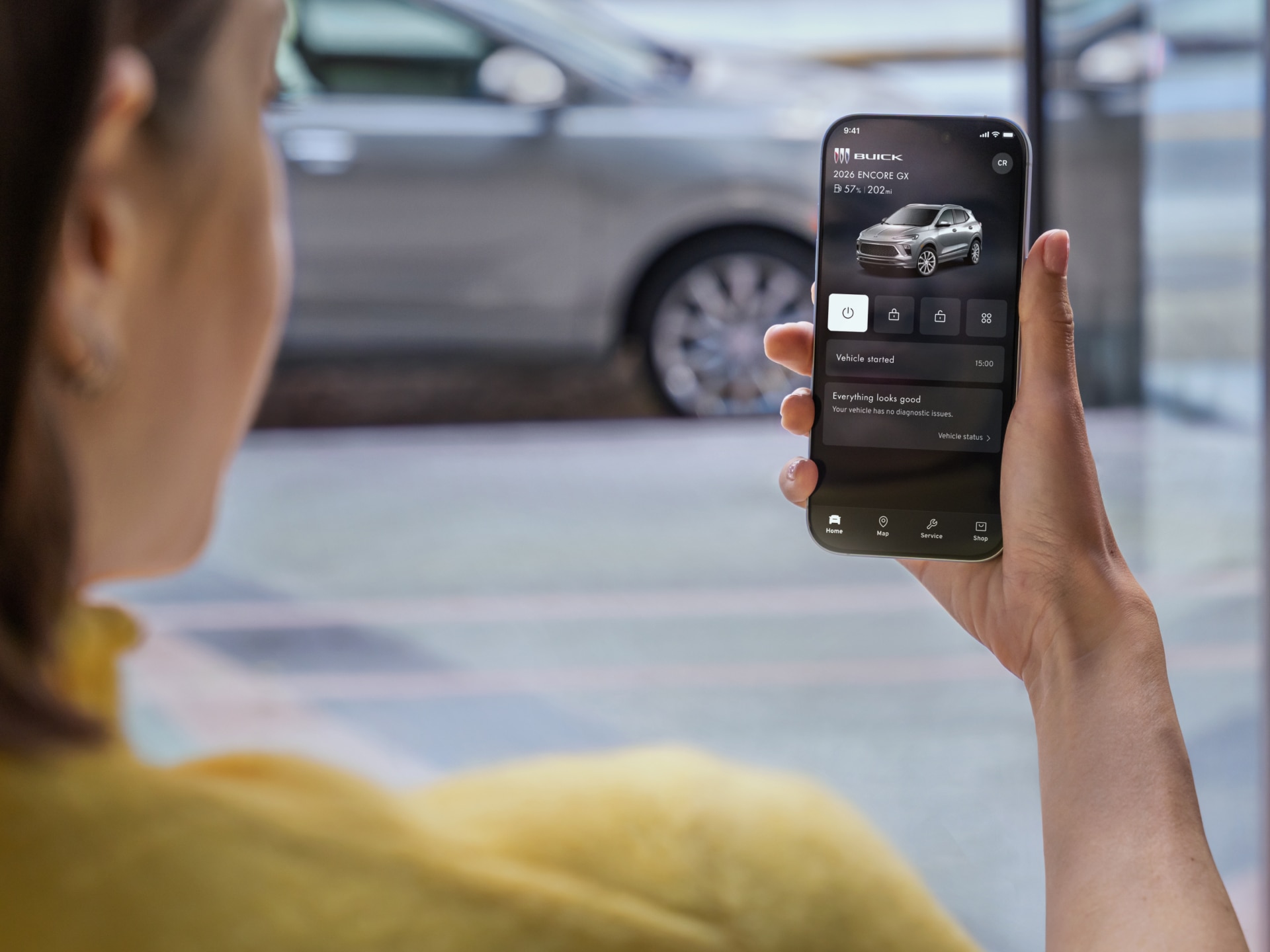  A Person Holding a Smartphone Displaying the Buick Encore GX in an App Showing Fuel Level with a Real Buick Vehicle in the Background