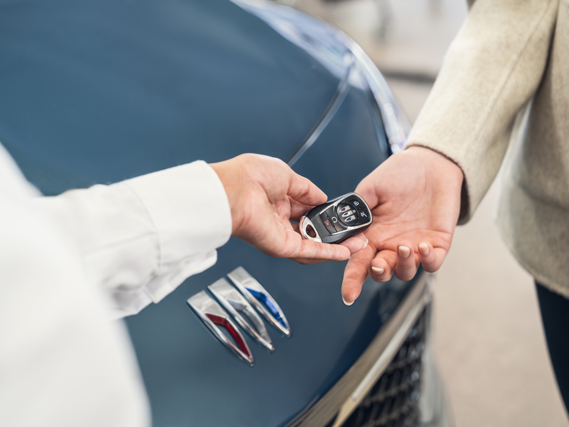 Buick key fob being handed over to another person with the Buick Logo on the hood of vehicle