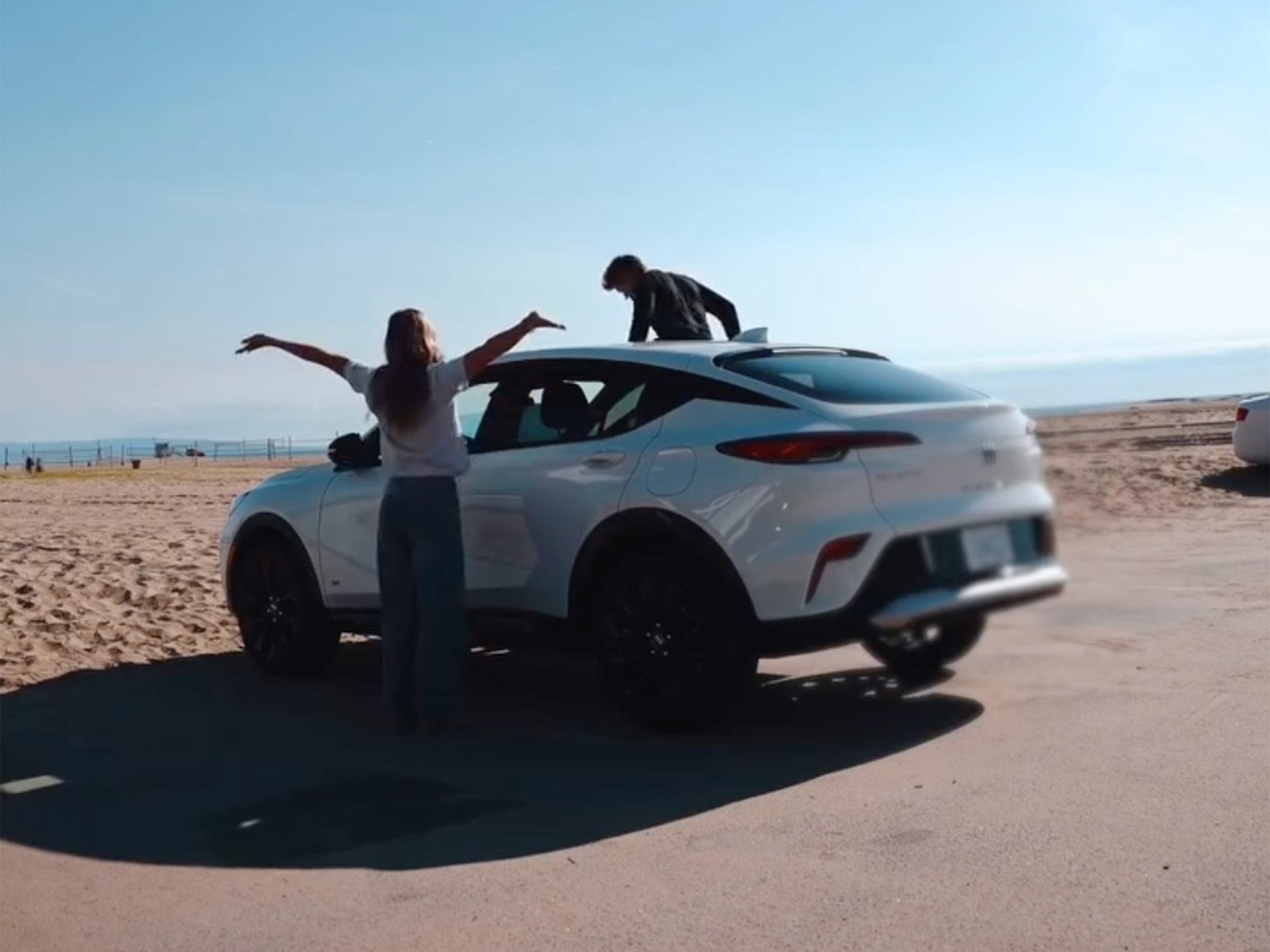 A White Buick Envista Parked on a Sandy Beach Road with a Woman Standing Beside it and a Man Climbing on the Roof 