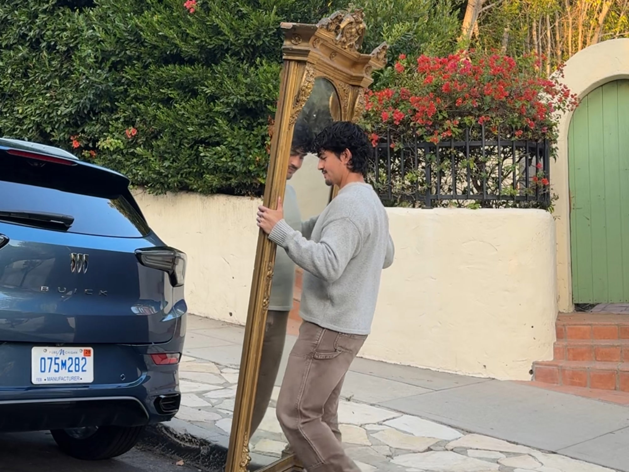 The Rear of a Dark Blue Buick SUV Parked on a Street as Marco Zamora Carries a Large Ornate Gold Framed Mirror Towards it