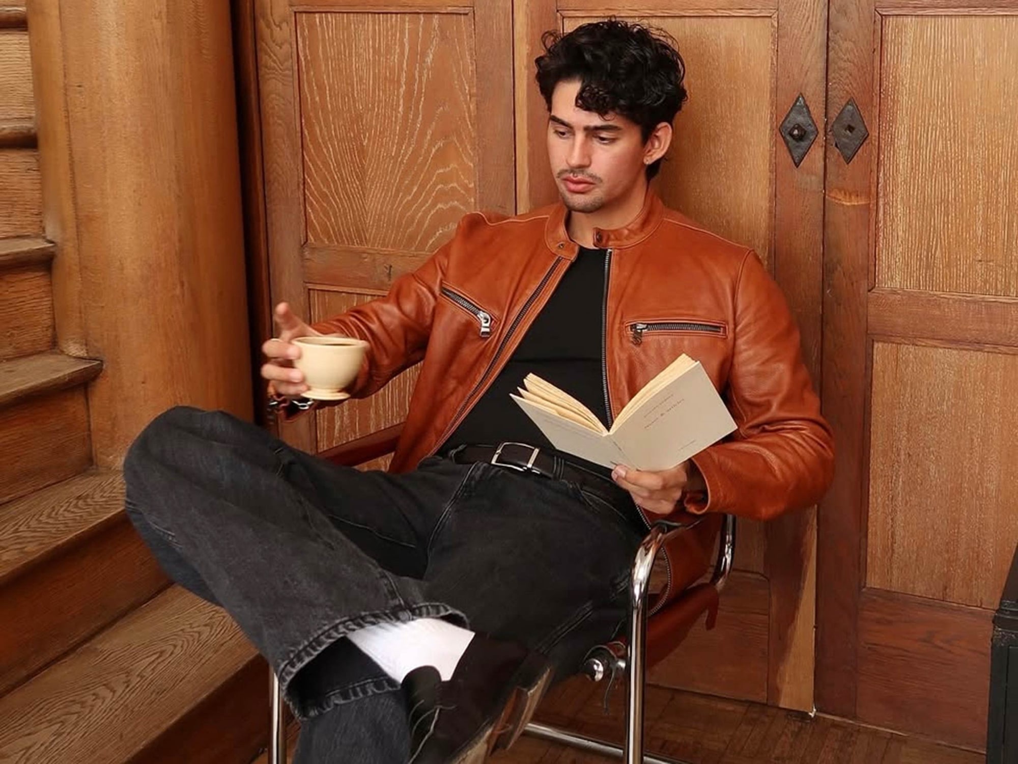 Marco Zamora Sitting In a Chair Reading a Book With a Brown Leather Jacket On