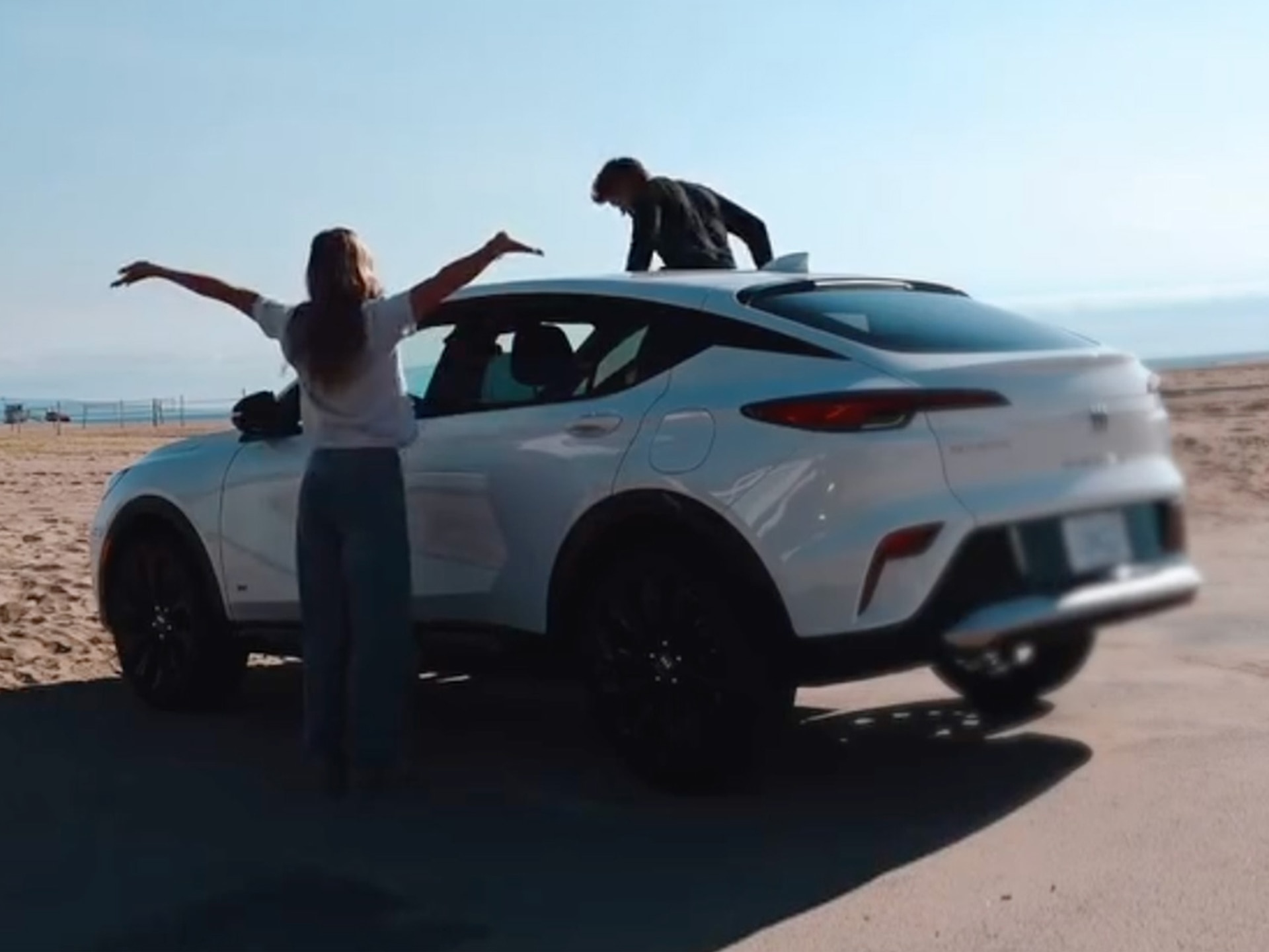 A White Buick Envista Parked on a Sandy Beach Road with a Woman Standing Beside it and a Man Climbing on the Roof