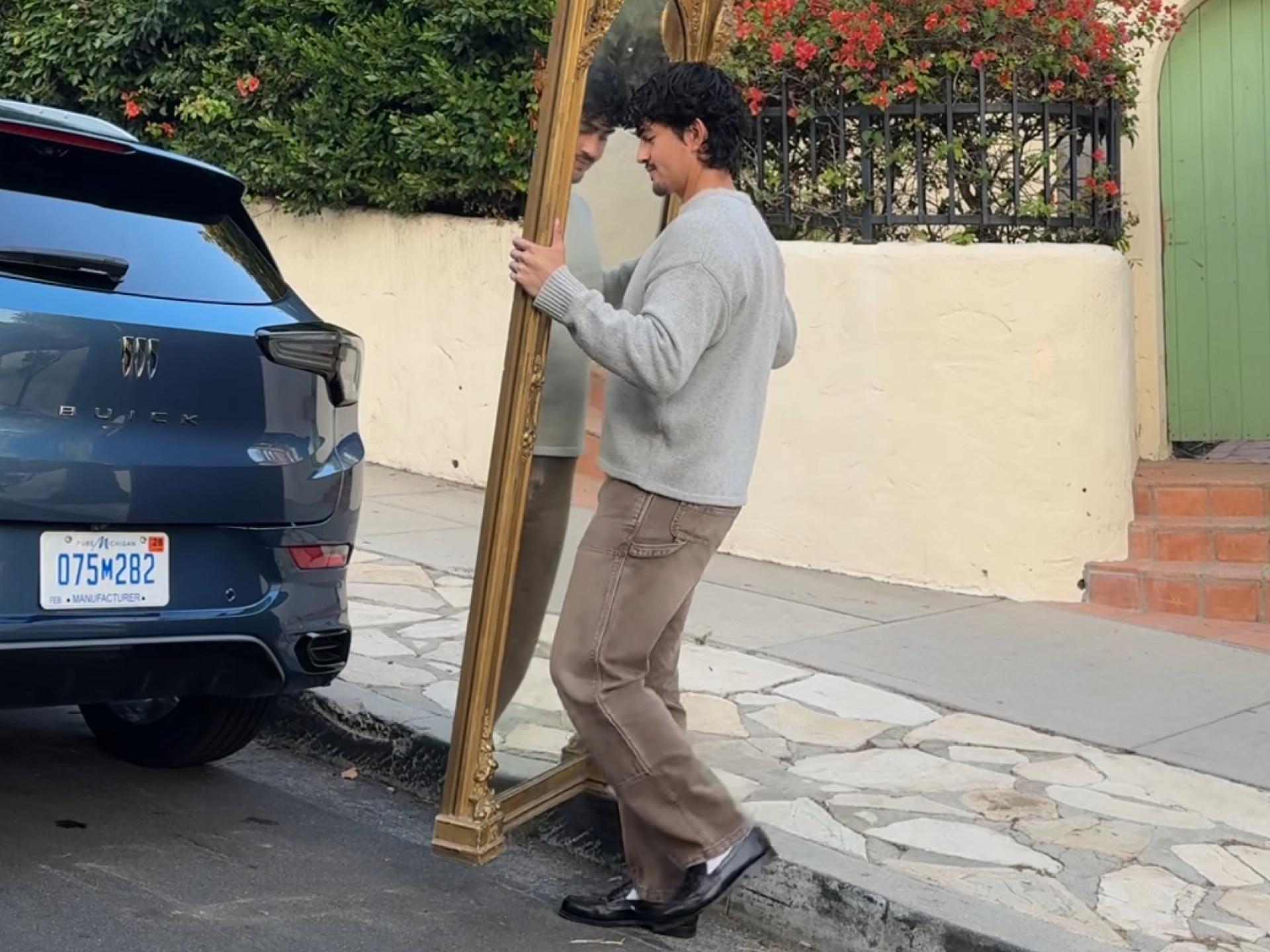 The Rear of a Dark Blue Buick SUV Parked on a Street as Marco Zamora Carries a Large Ornate Gold Framed Mirror Towards it