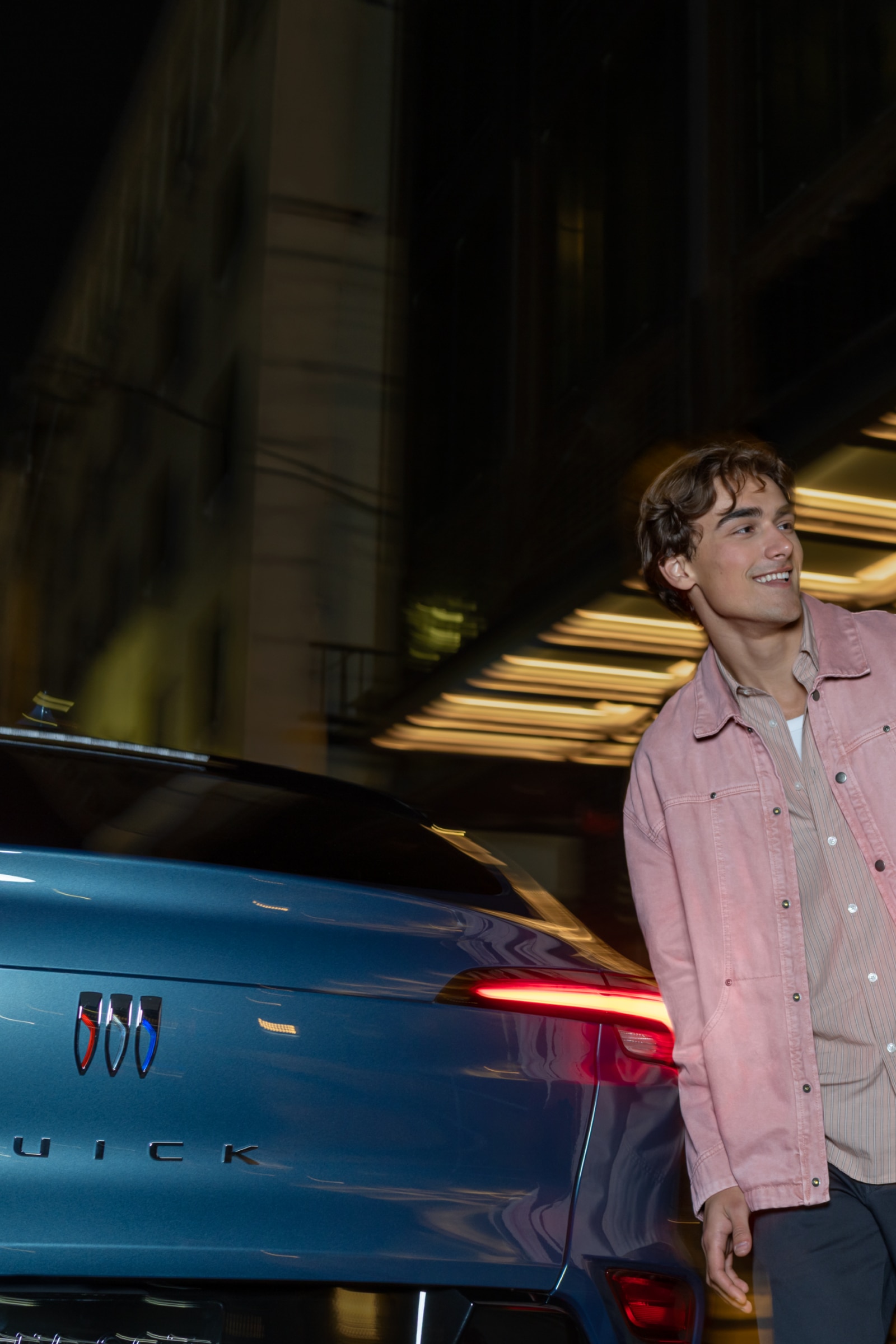 A Young Couple Walking Hand in Hand Past a Blue Buick SUV at Night with City Lights in the Background
