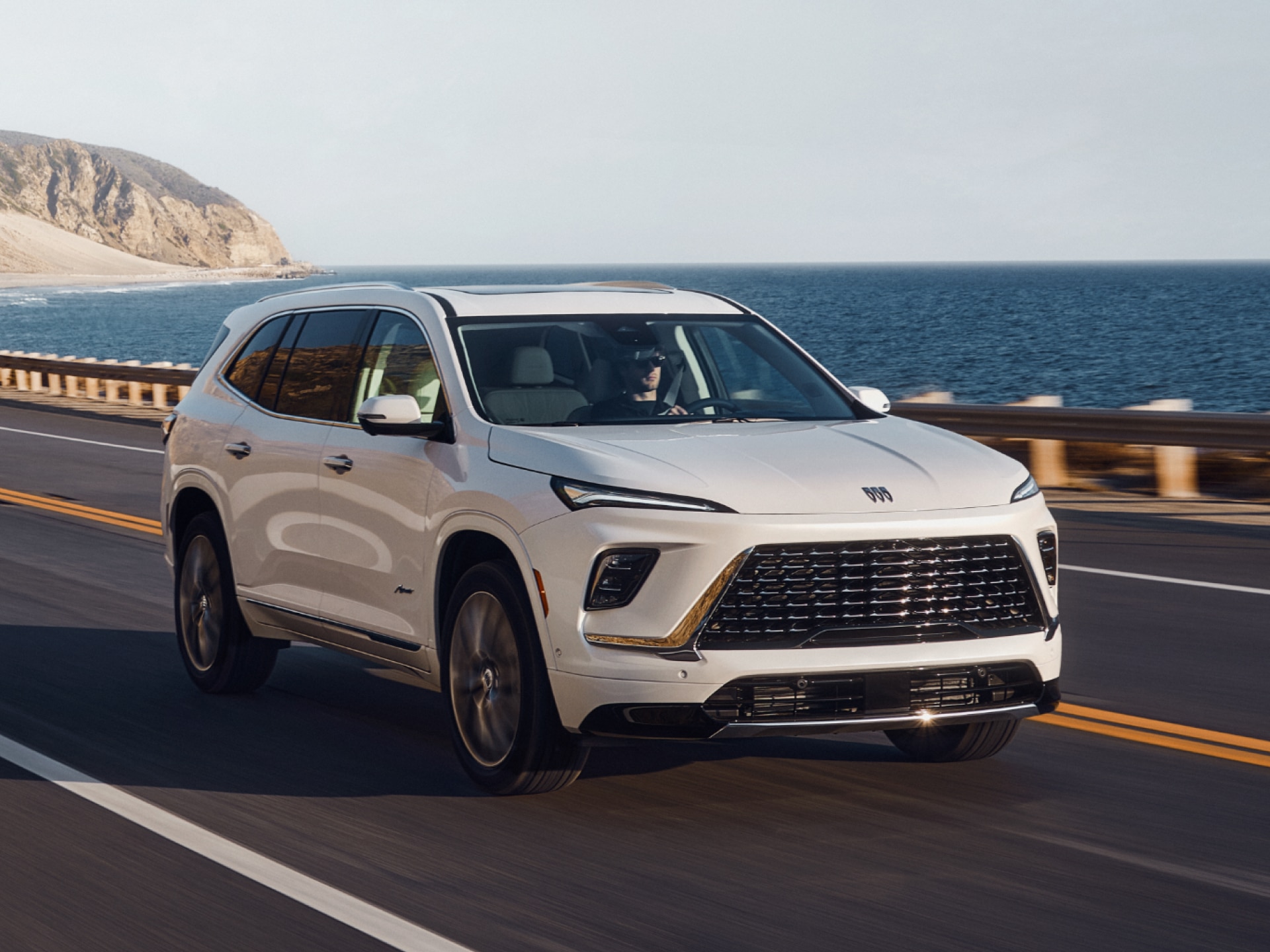 A White Buick Enclave SUV Driving on a Coastal Road Overlooking the Ocean with a Distant Rocky Cliff Under a Clear Sky