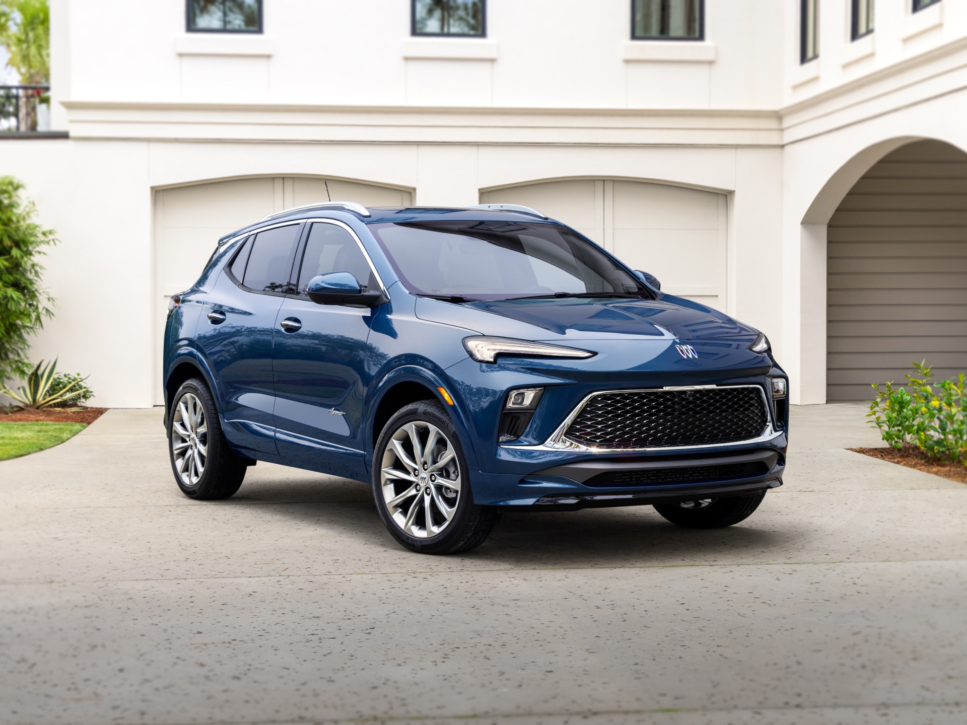 A Front Quarter View of a Shiny Dark Blue Buick Envista SUV with Silver Wheels Parked on a Paved Driveway in Front of a White House with Garage Doors
