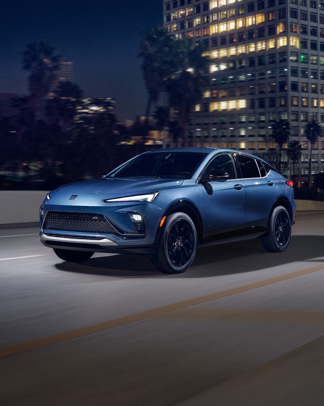 A Front Three-Quarters View of a Blue Buick Envista ST Driving at Night Along a Lit City Street