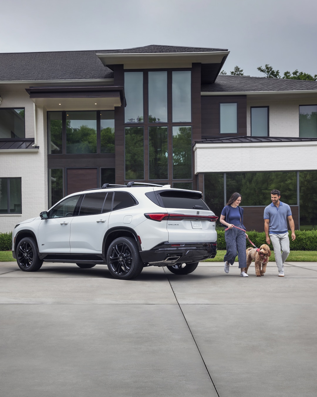 A White Buick SUV with Black Wheels is Parked on a Driveway with a Couple and Their Dog Walking Away from It