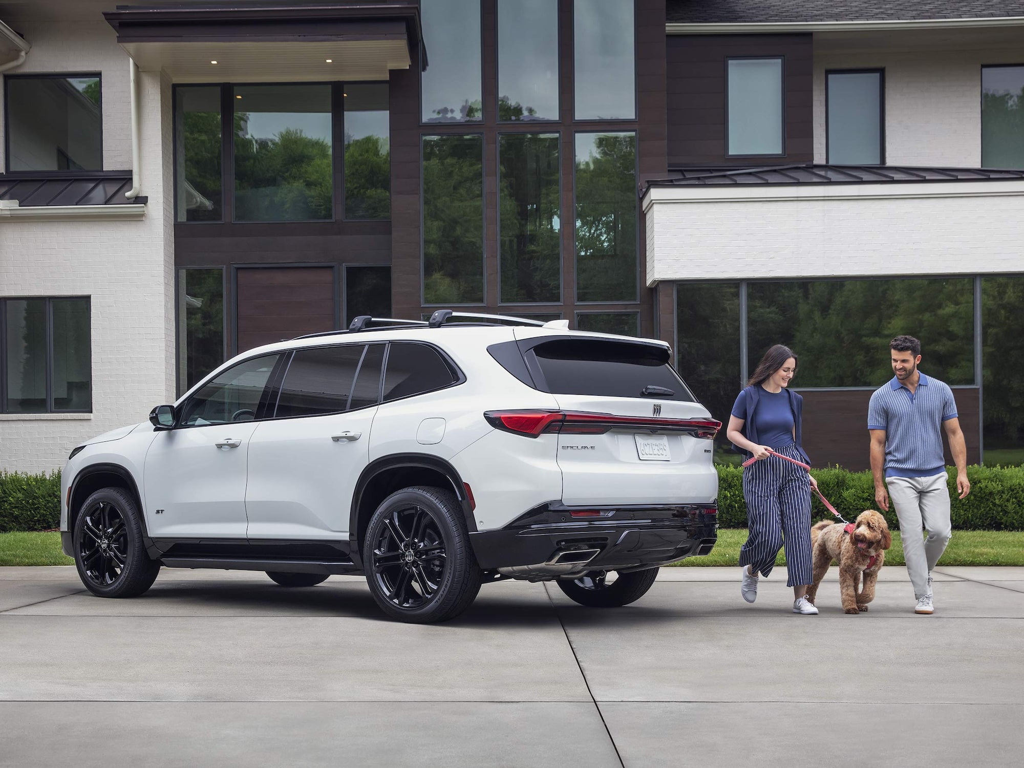 A White Buick SUV with Black Wheels is Parked on a Driveway with a Couple and Their Dog Walking Away From It