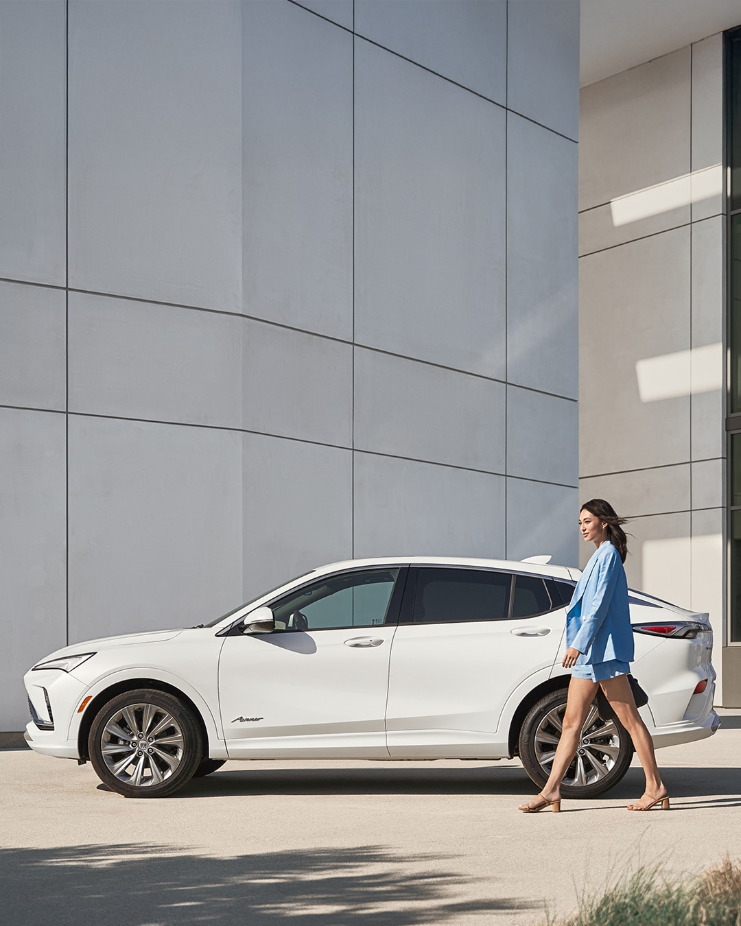 A White Buick Envision Avenir SUV Is Parked as a Woman in a Light Blue Blazer Walks by in Front of a Modern Gray Building