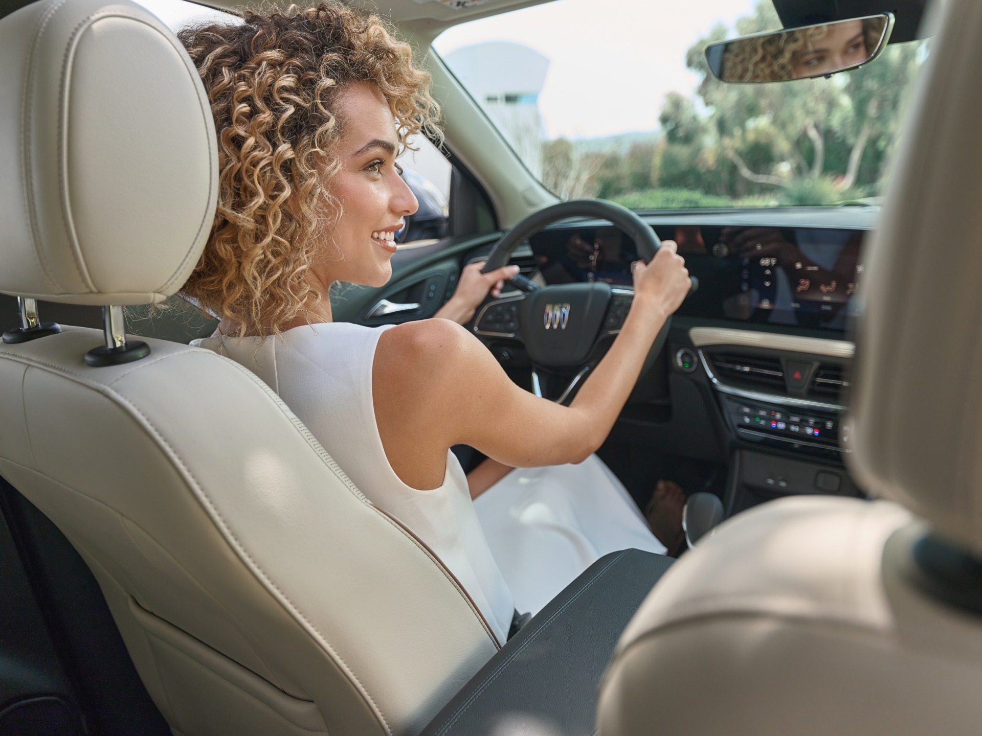 A Smiling Woman with Curly Blond Hair Driving a Buick Envision with  Light Colored Seats and a Digital Dashboard