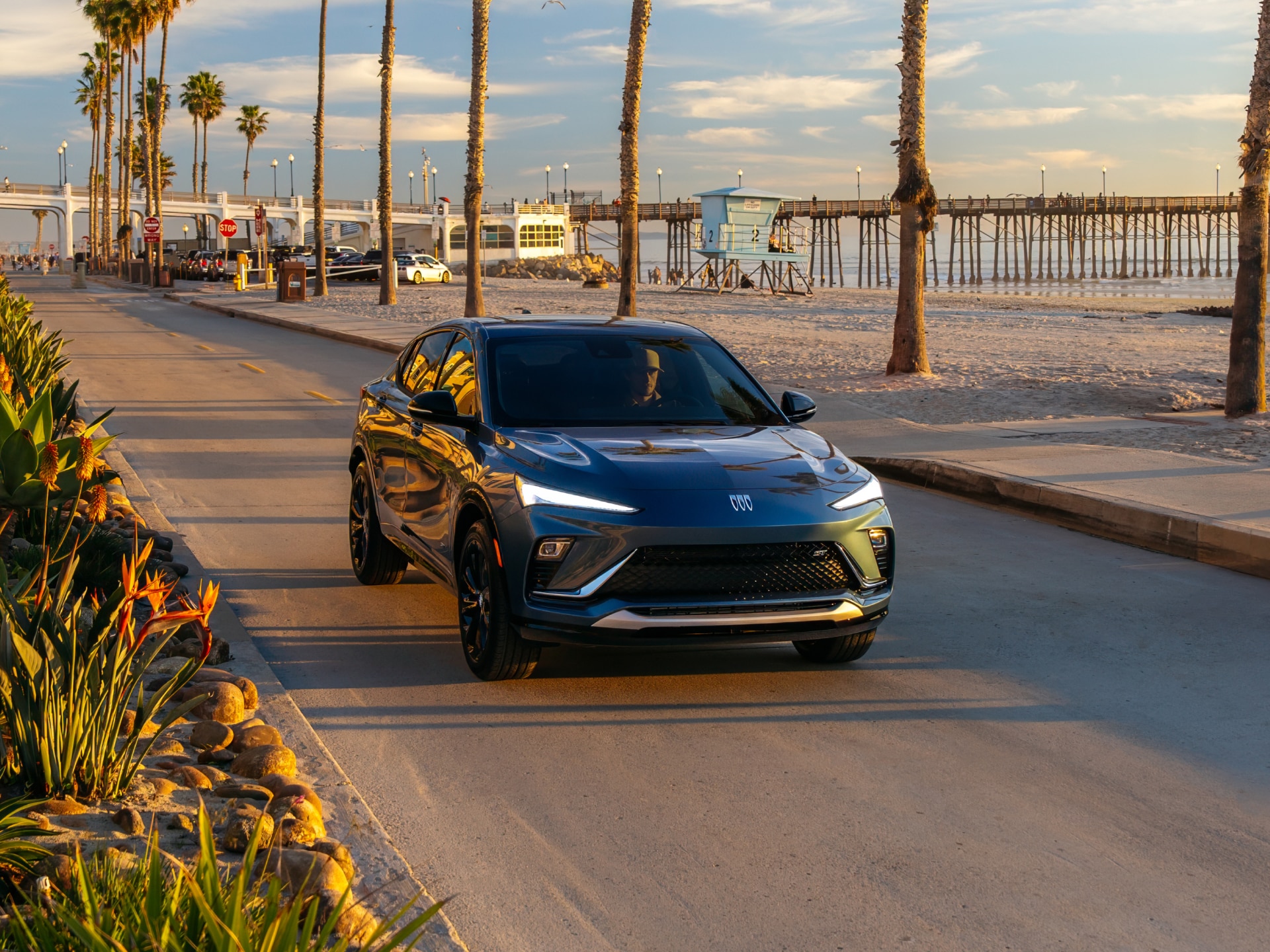 A Dark Blue Buick Envista Drives Along a Coastal Road with Palm Trees and a Pier in the Background
