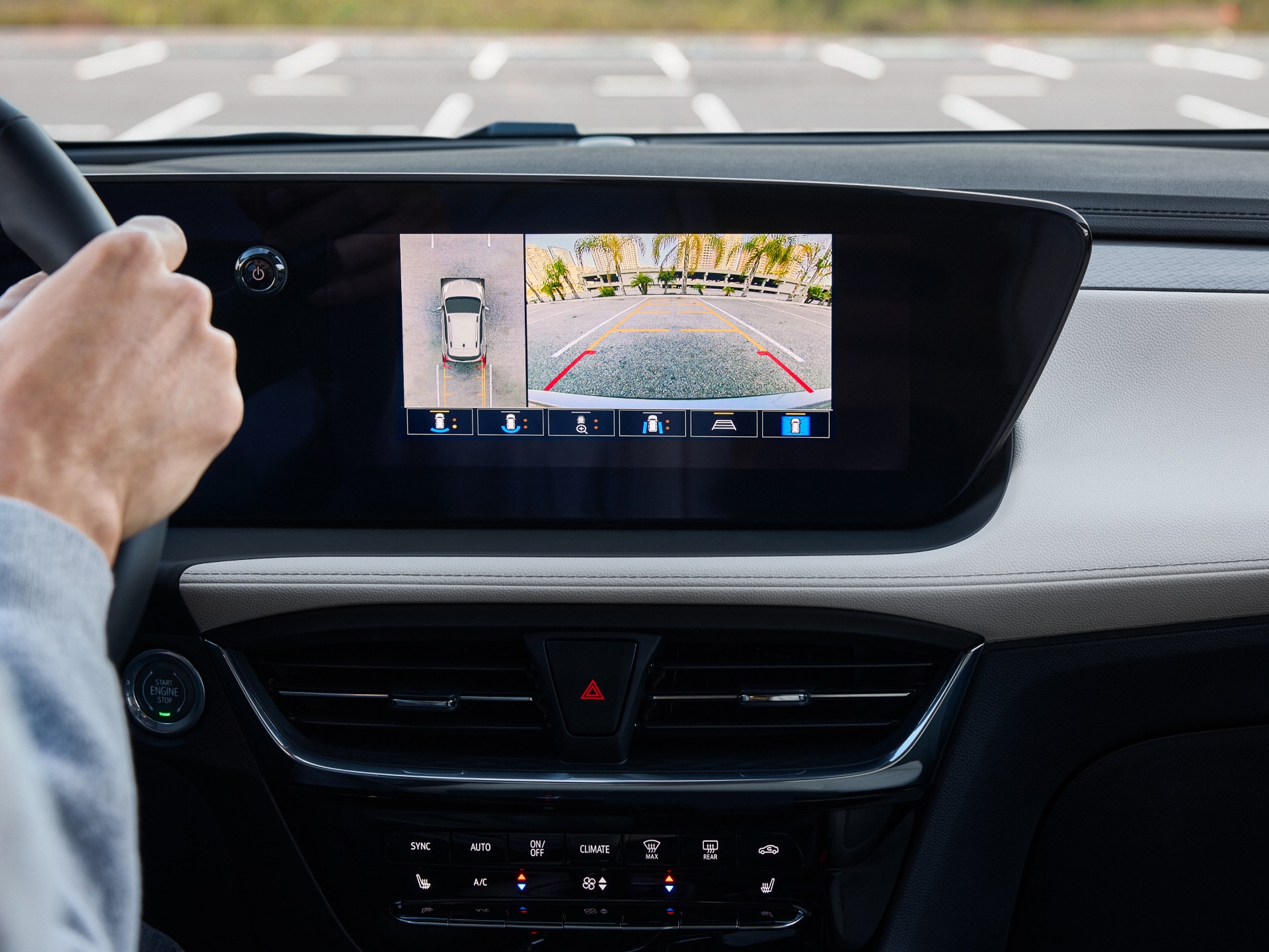 A Driver's Hands on a Buick Steering Wheel with the Infotainment Screen Showing a 360-Degree Camera View and Rear Parking Assist plus Climate Controls