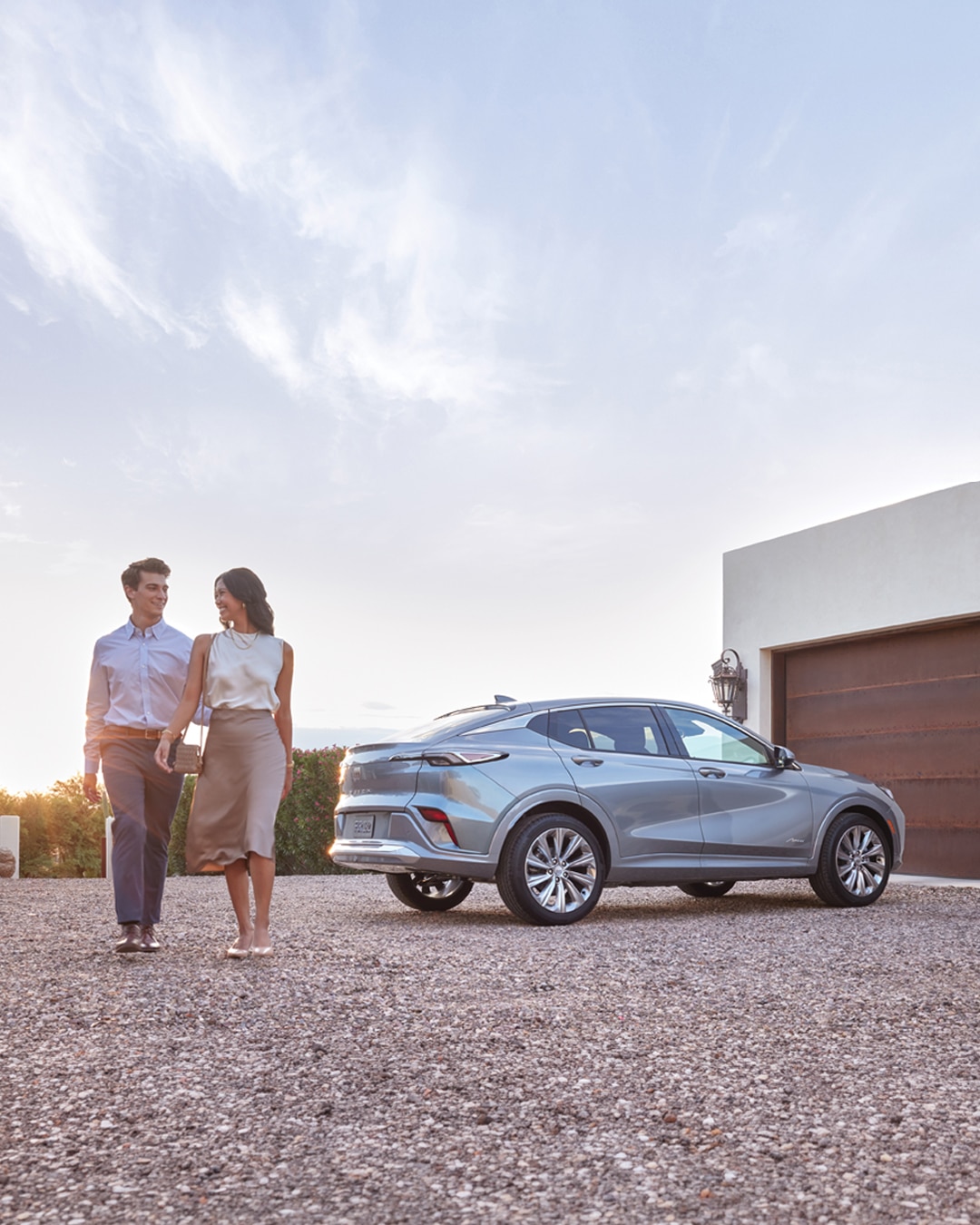 A Couple Walks Beside a Sleek Light Gray Buick SUV Parked Outside a Modern Home with a Garage During Sunset