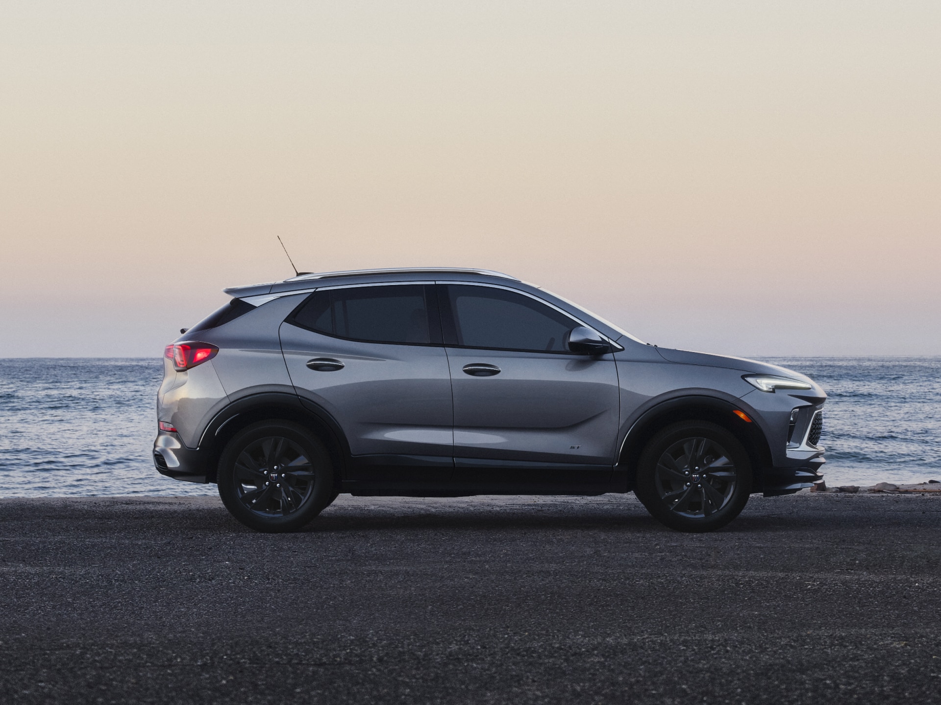 A Side Profile View of a Gray Buick Encore GX SUV Parked on the Asphalt Road by the Ocean at Sunset