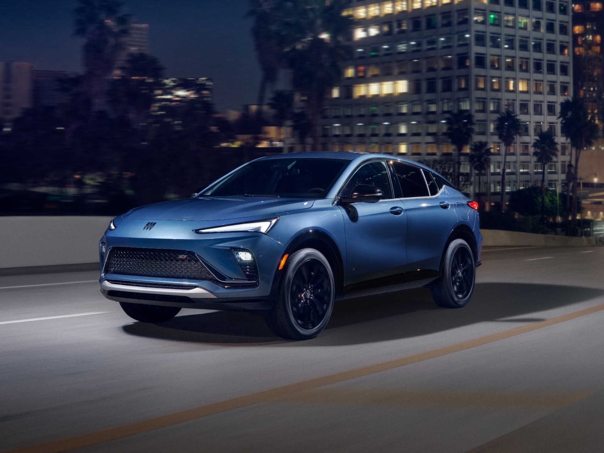 A Blue Buick Envista SUV Driving on a City Street at Night with Illuminated Buildings in the Background