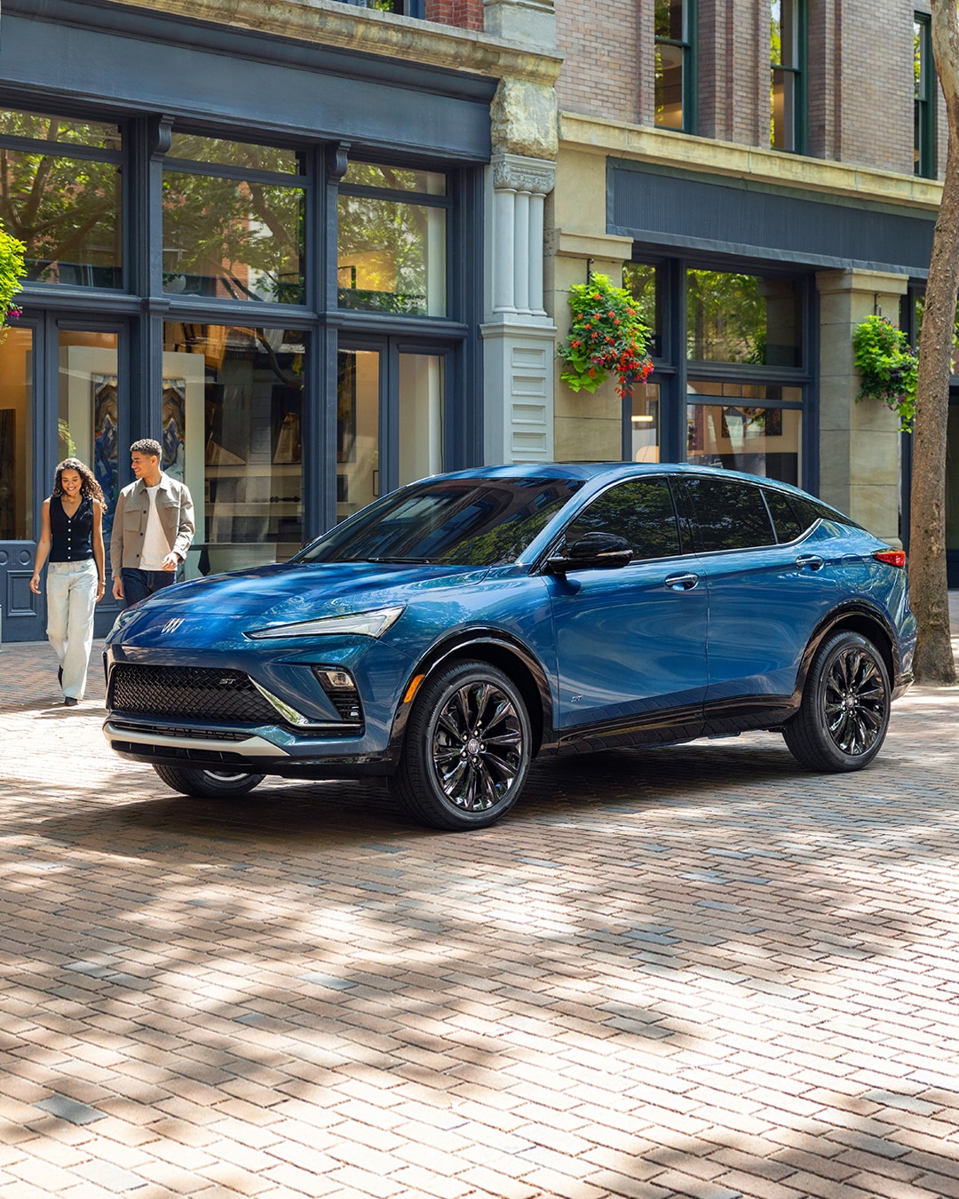 A Brilliant Blue Buick Envista ST SUV Parked on a Brick Sidewalk as Two People Walk Past a Storefront in the Background