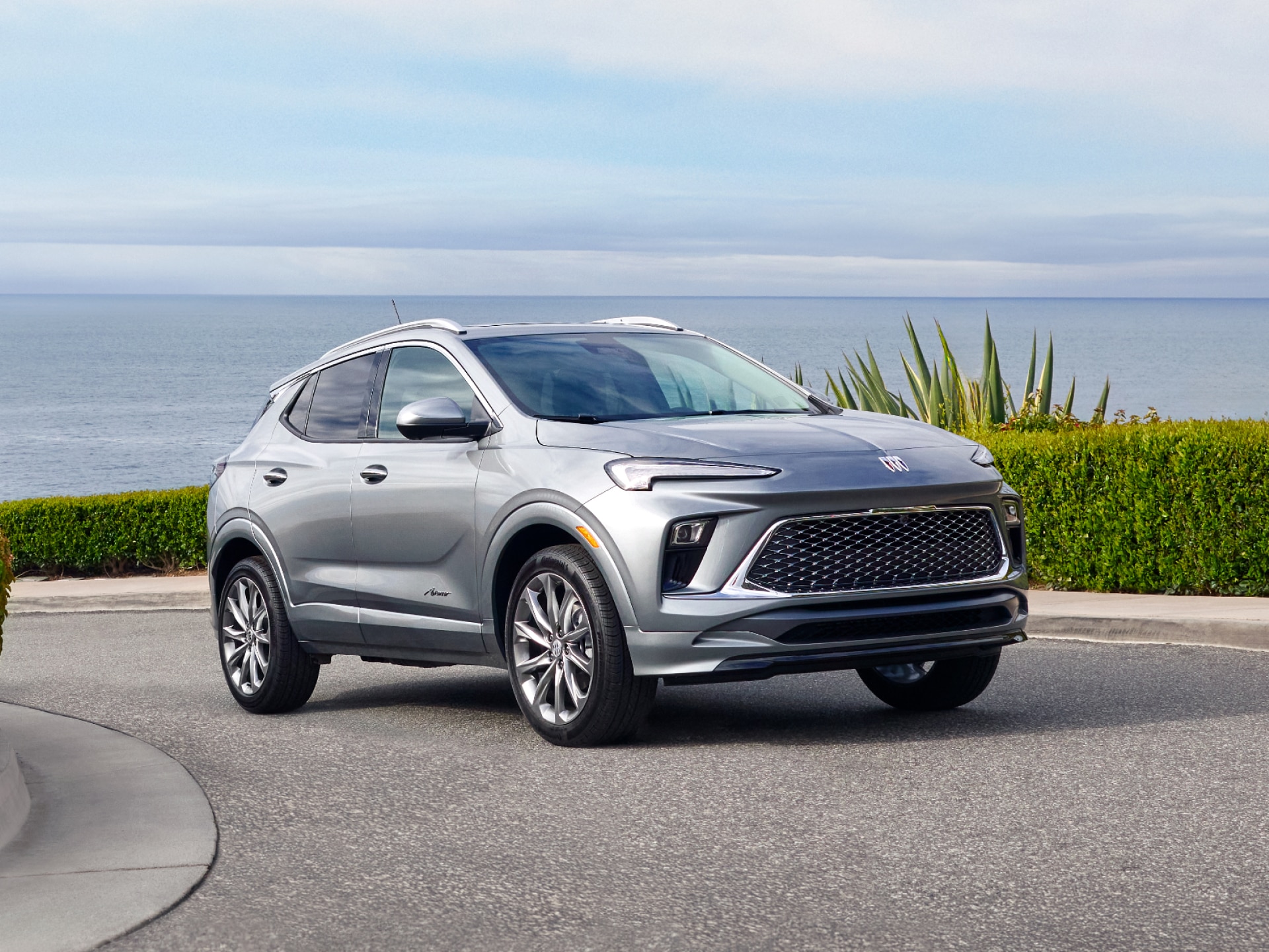 A Silver Buick Envista Parked on an Asphalt Road with the Ocean and Green Plants in the Background