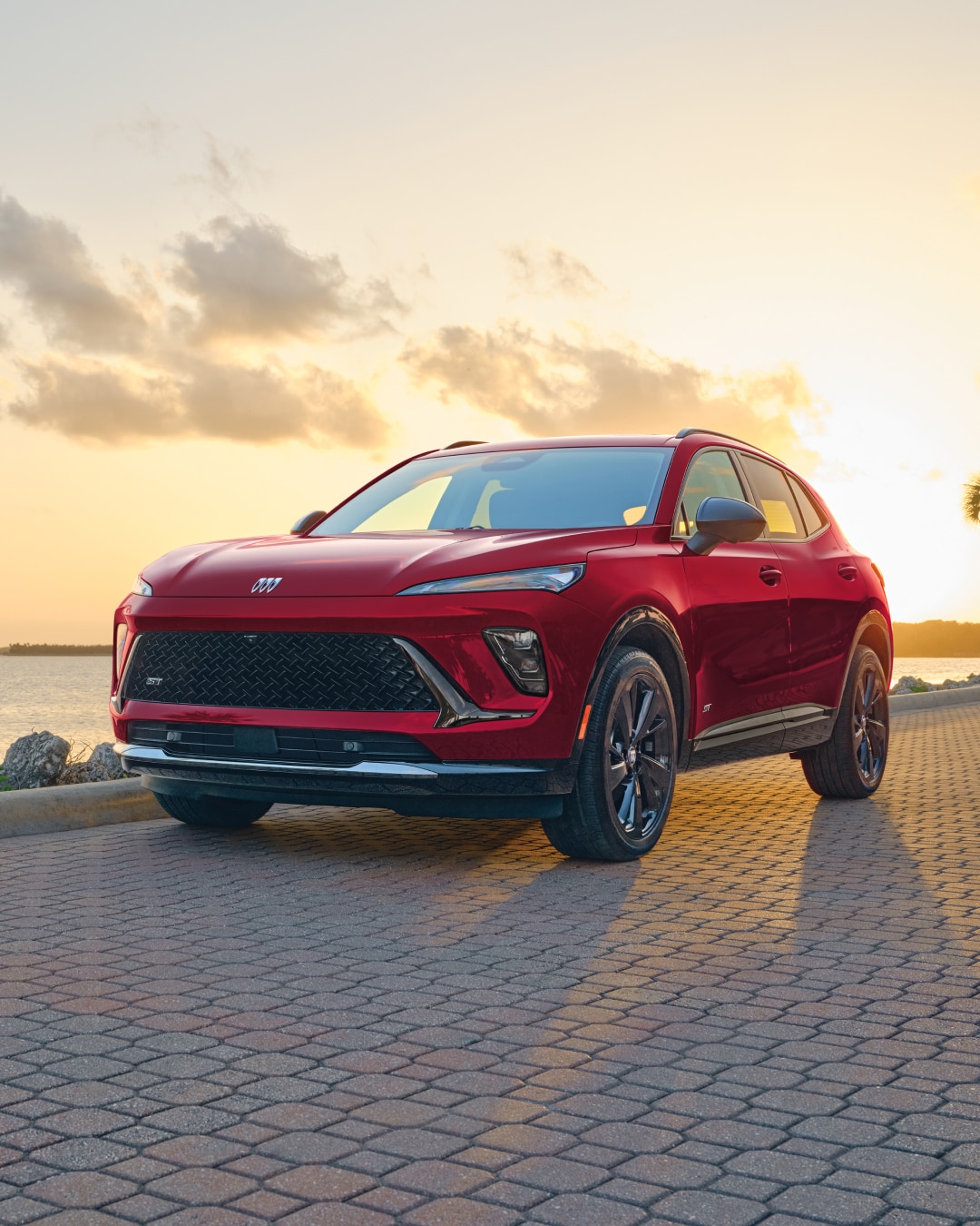 A Red Buick Envista ST Parked on a Patterned Brick Road with a Sunset Sky and Water in the Background