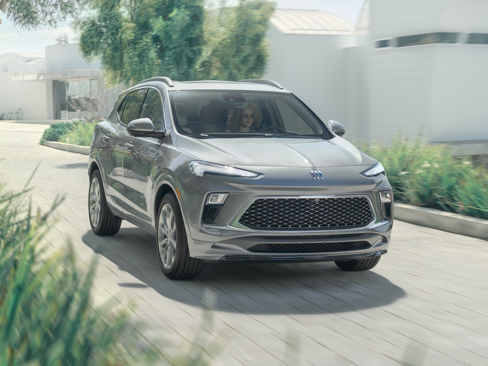 A Gray 2026 Buick Encore GX SUV Parked in a Driveway of a Modern House with Green Foliage 