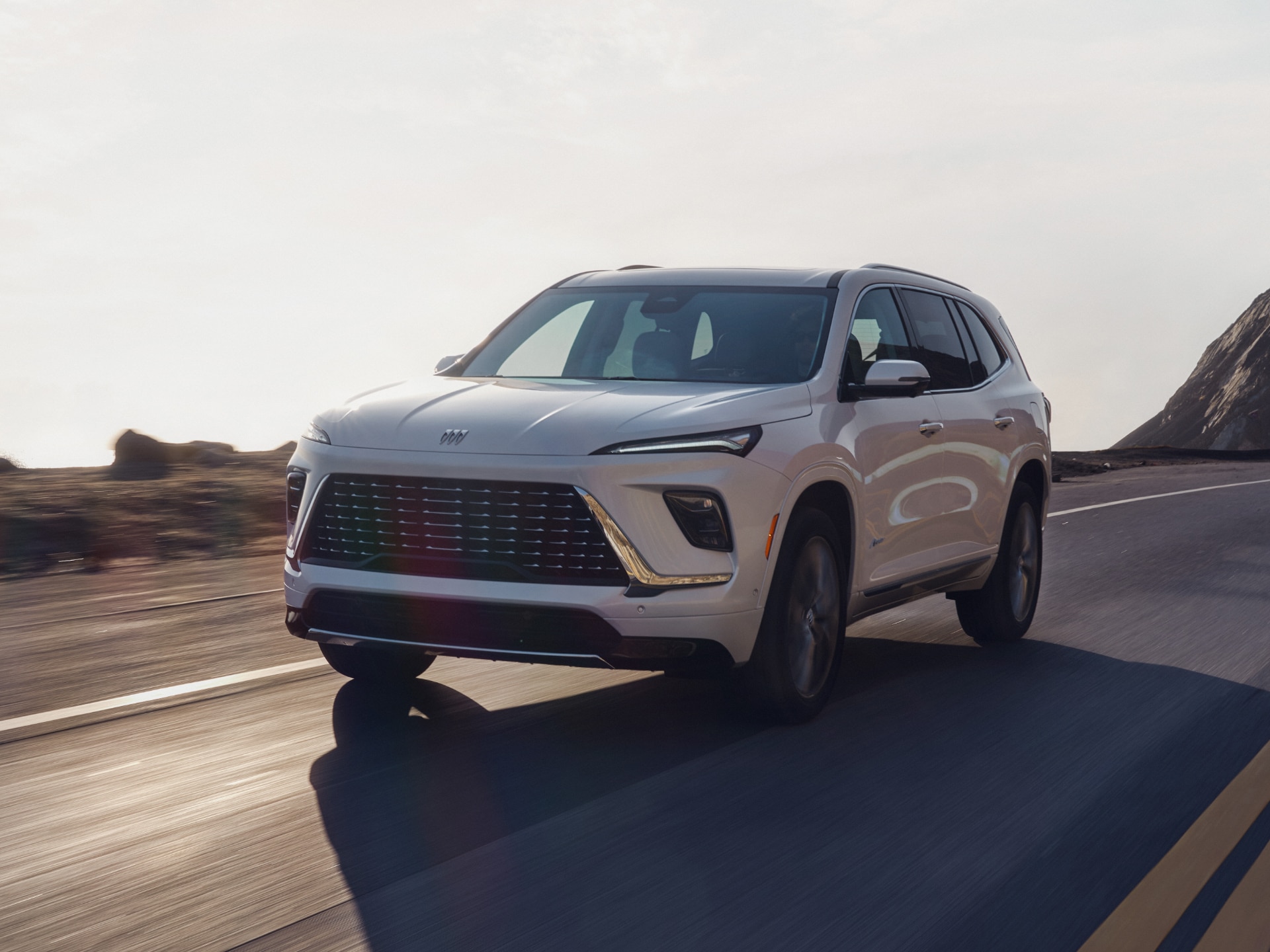 A white Buick Enclave SUV driving along a winding road at sunset with mountains in the background.