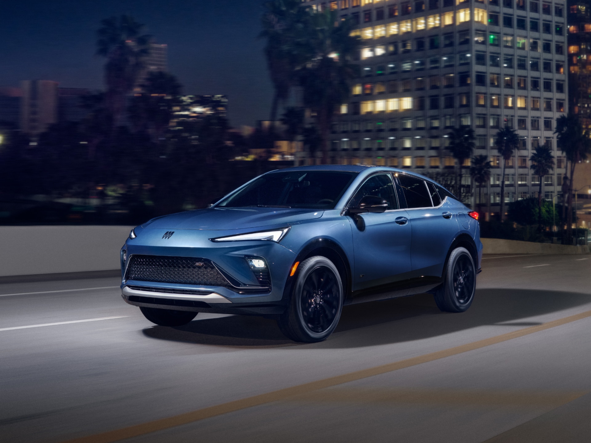 A blue Buick Envista SUV driving on a city street at night with illuminated buildings in the background.