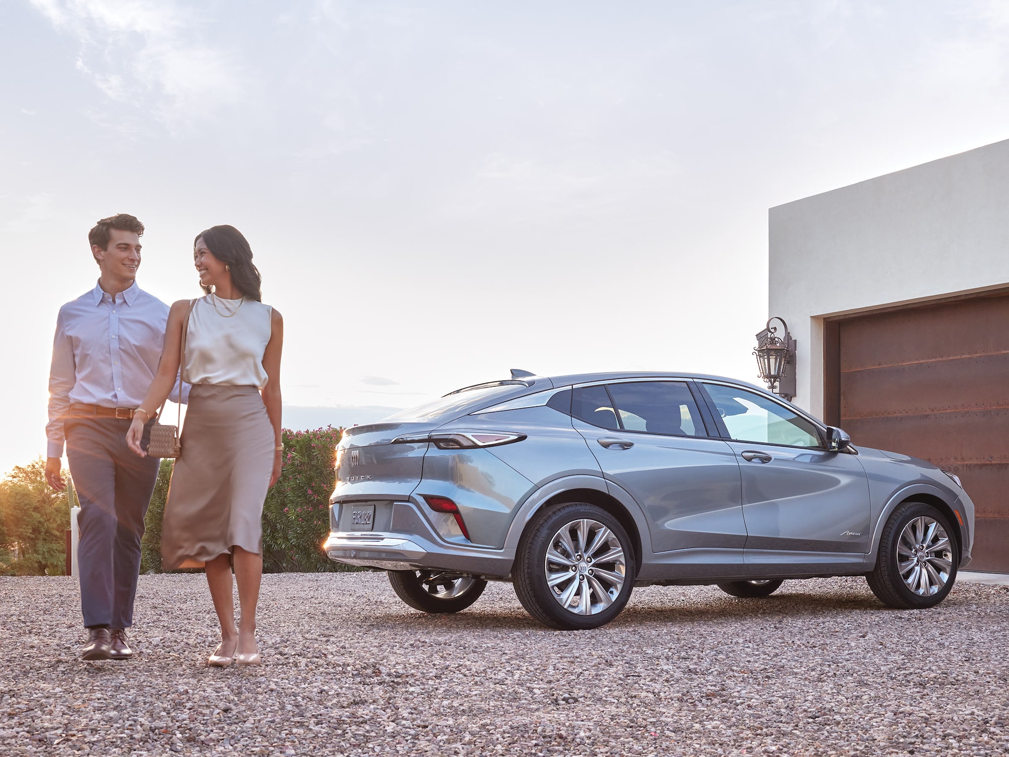A Couple Walks Beside a Sleek Light Gray Buick SUV Parked Outside a Modern Home with a Garage During Sunset