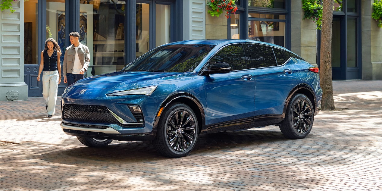 A Brilliant Blue Buick Envista ST SUV Parked on a Brick Sidewalk as Two People Walk Past a Storefront in the Background