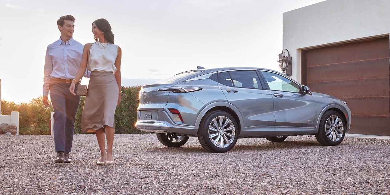 A Couple Walks Beside a Sleek Light Gray Buick SUV Parked Outside a Modern Home with a Garage During Sunset