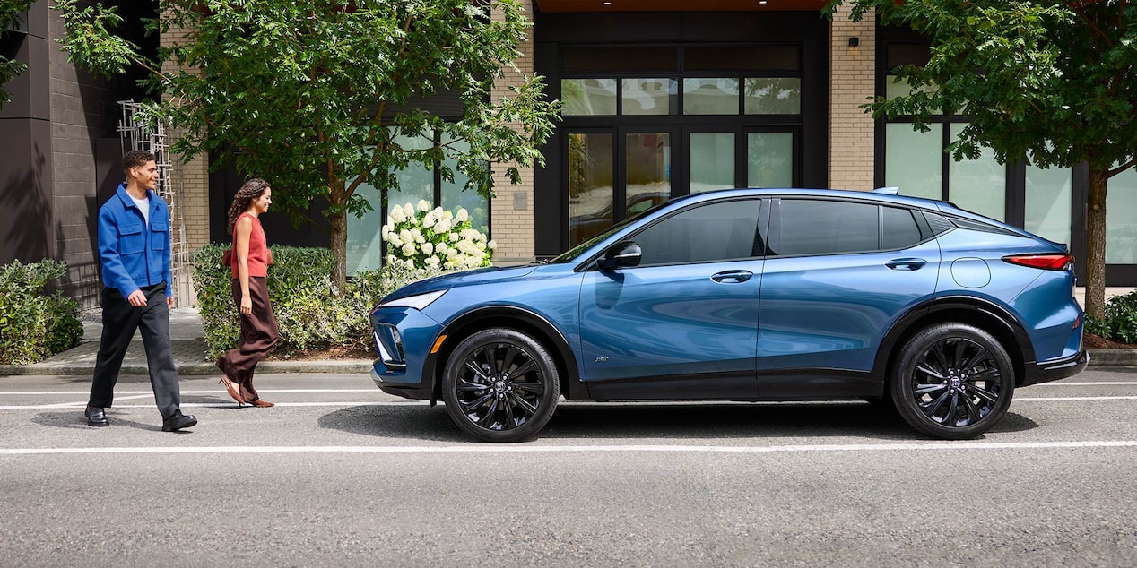 A Metallic Blue Buick Envista Parked on a City Street with a Couple Walking in Front of it and Green Foliage in the Background