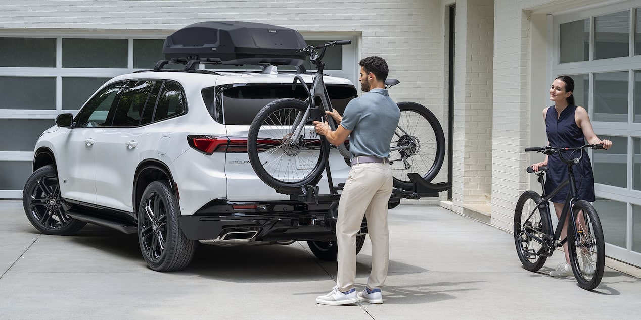 A White Buick SUV with a Roof Rack and Bike Carrier Is Parked in a Driveway a Man Loads a Bicycle onto the Rear Rack While a Woman Stands Nearby with Another Bicycle