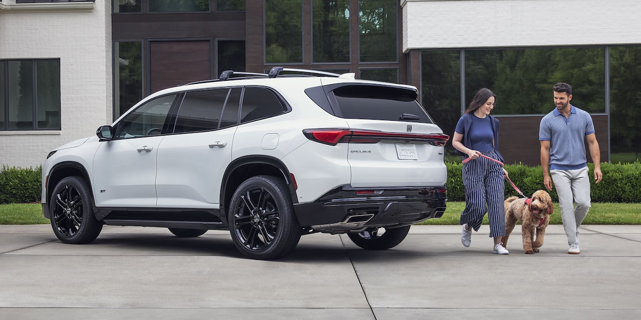 A White Buick SUV with Black Wheels is Parked on a Driveway with a Couple and Their Dog Walking Away From It