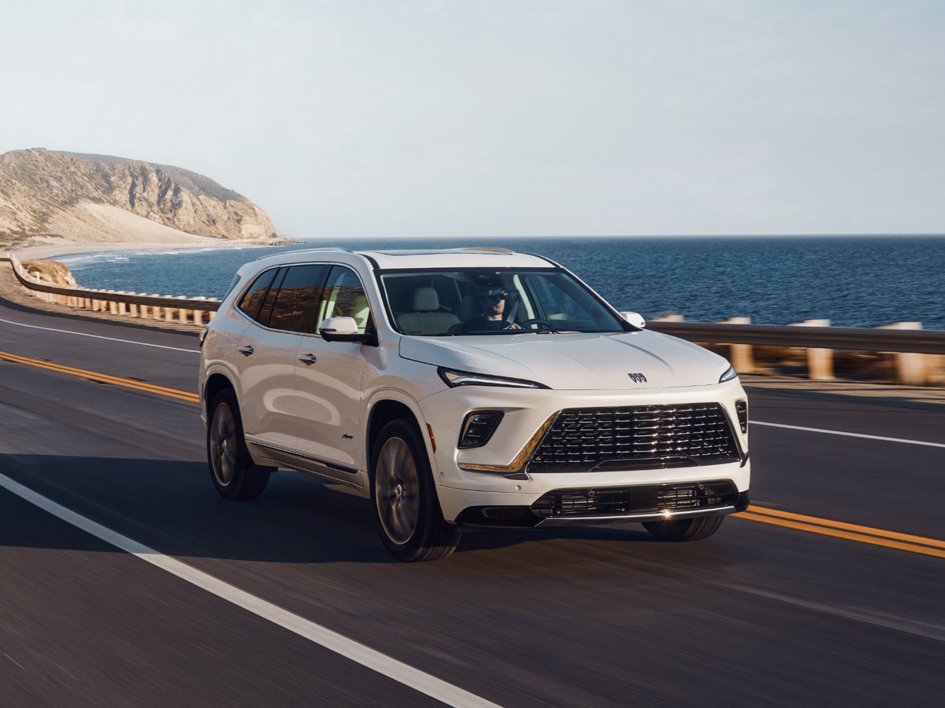 A Three-Quarters Front View of a Man Driving a White Buick SUV Along the Coast on a Sunny Day