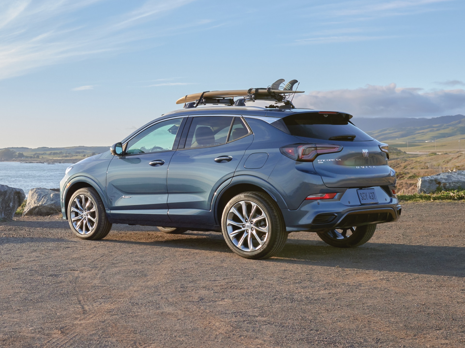 Rear Three-Quarters View of a Buick Encore GX Avenir Luxury SUV Parked Overlooking a Large Body of Water with a Surfboard Attached to the Top of the Vehicle
