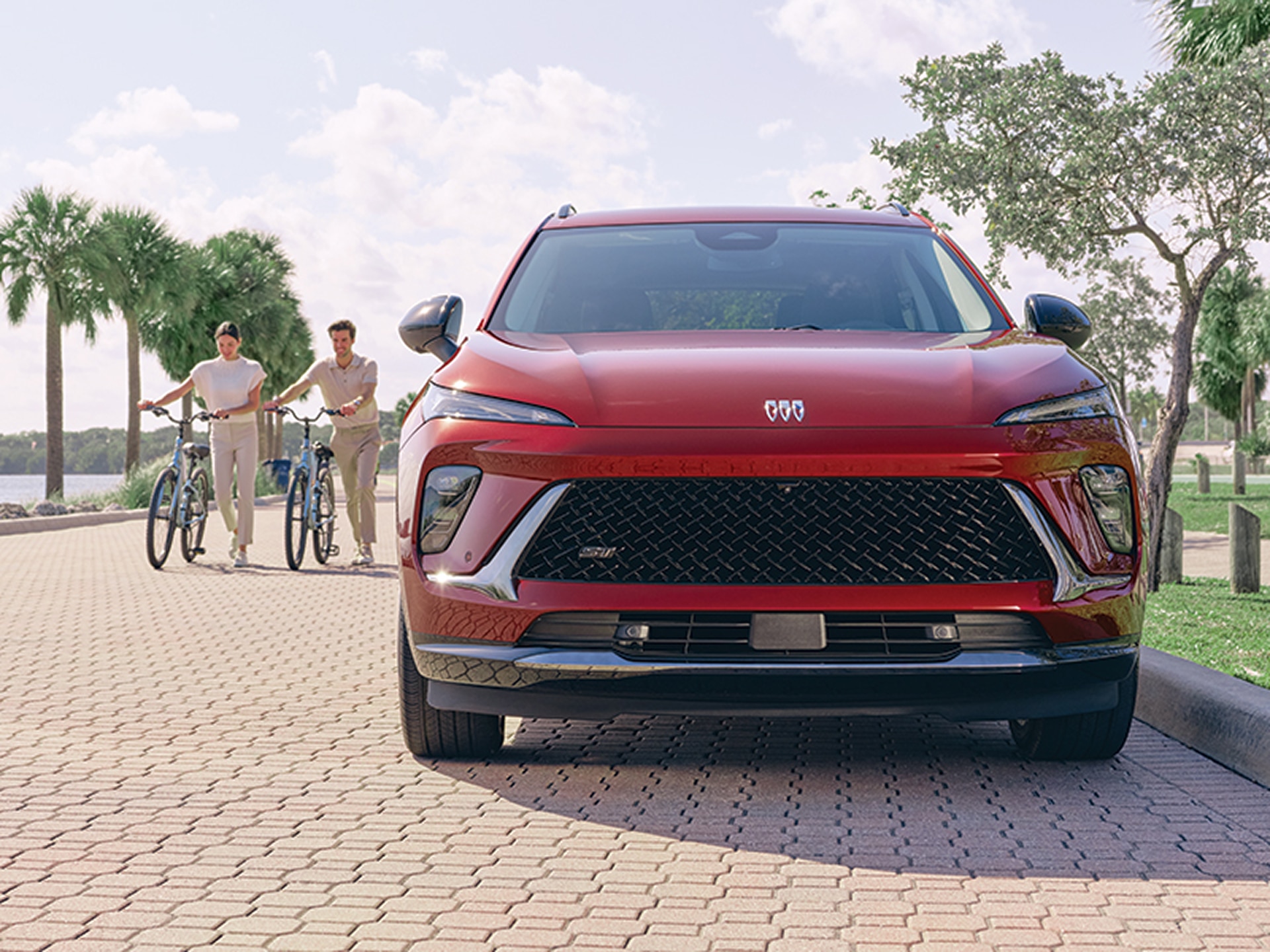 Front View of a Red Buick Envision Compact SUV Parked on a Tropical Street with Two Bikers Passing by