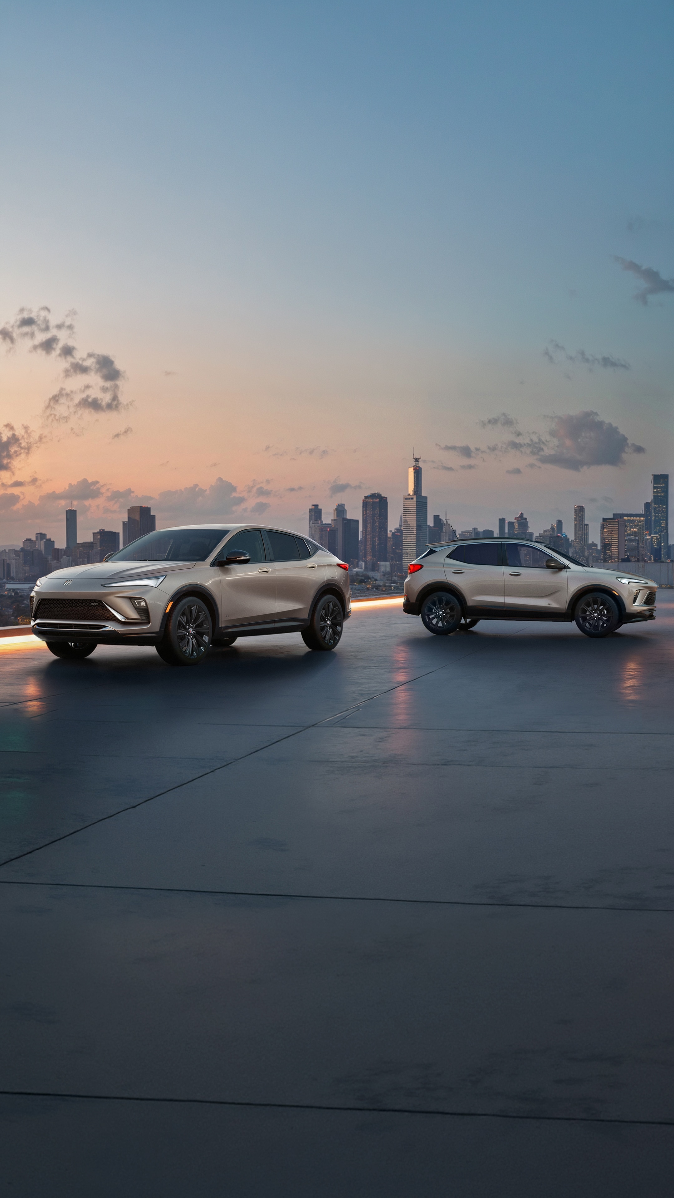 The 2026 Envista Sport Touring SUV and the 2026 Encore GX Sports Touring SUV both in the Color Ceramic Parked on a Parking Garage Rooftop Overlooking a City Skyline