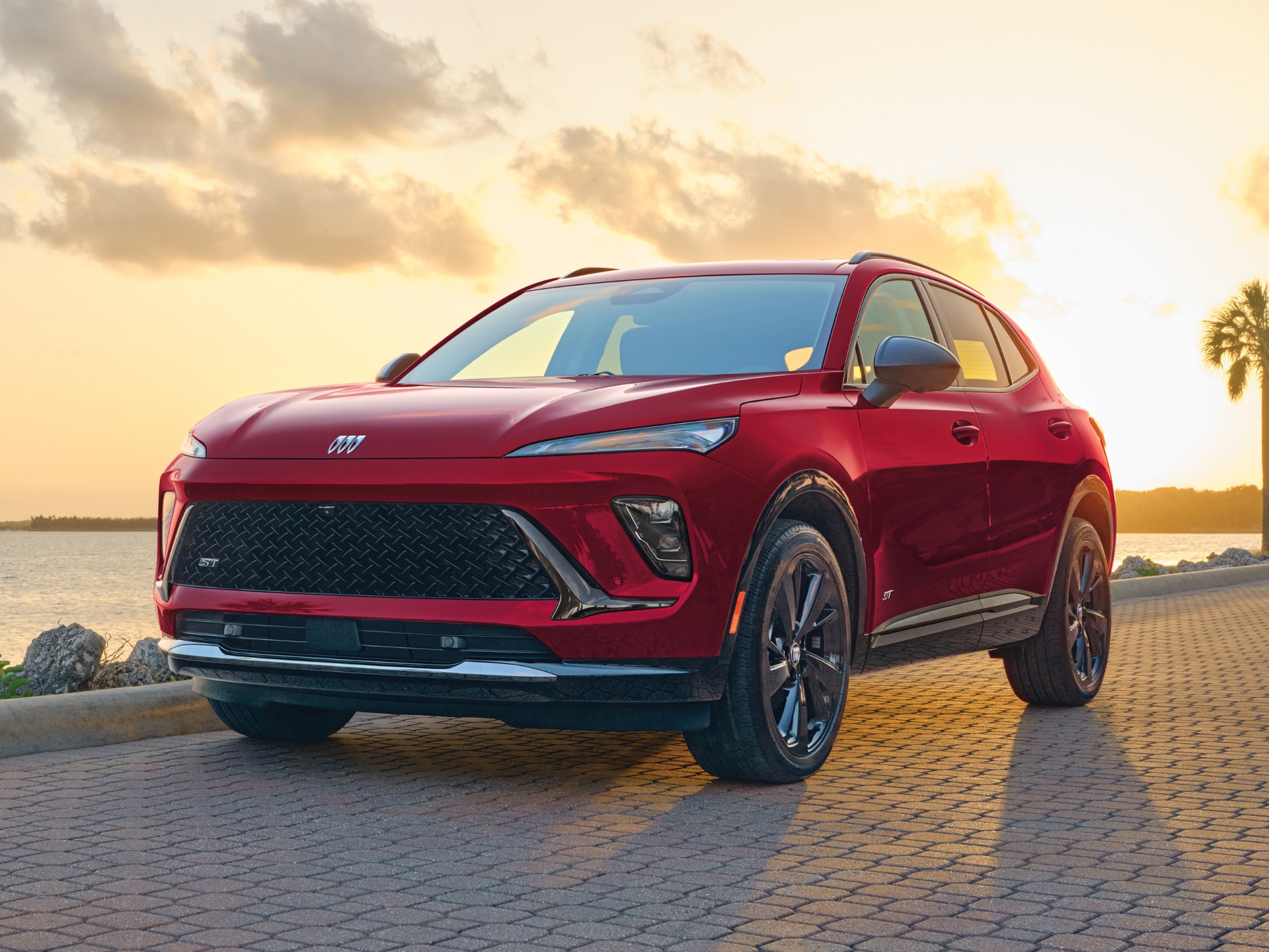 A Red Buick Envista ST Parked on a Patterned Brick Road with a Sunset Sky and Water in the Background