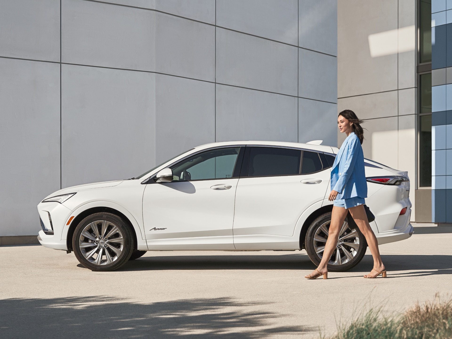 A White Buick Envision Avenir SUV is Parked as a Woman in a Light Blue Blazer Walks by in Front of a Modern Gray Building