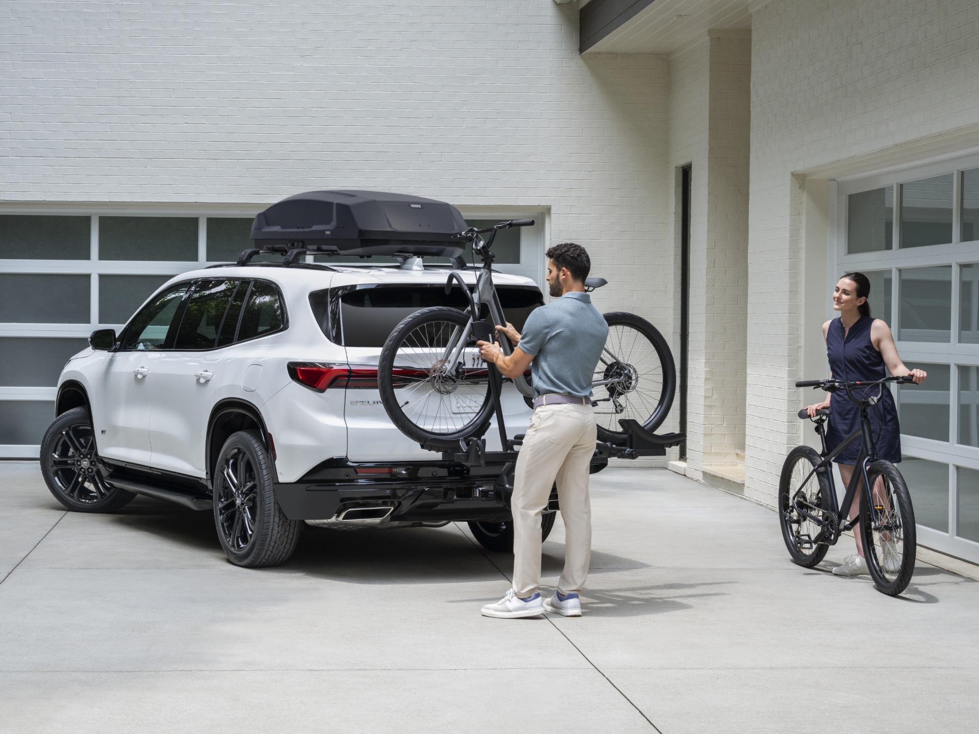 A White Buick SUV with a Roof Rack and  Bike Carrier is Parked in a Driveway a Man Loads a Bicycle onto the Rear Rack While a Woman Stands Nearby with Another Bicycle