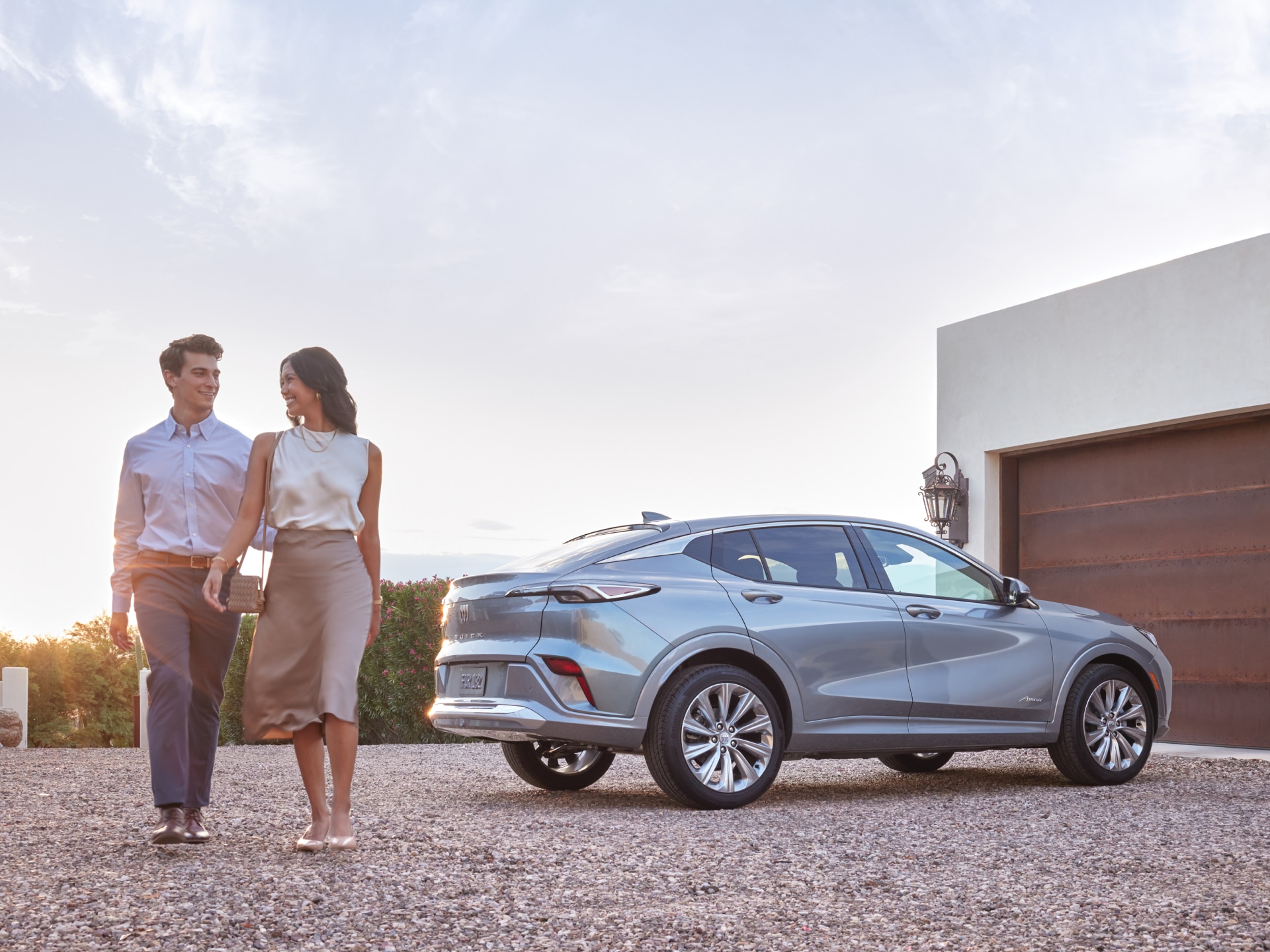 A Couple Walks Beside a Sleek Light Gray Buick SUV Parked Outside a Modern Home with a Garage During Sunset
