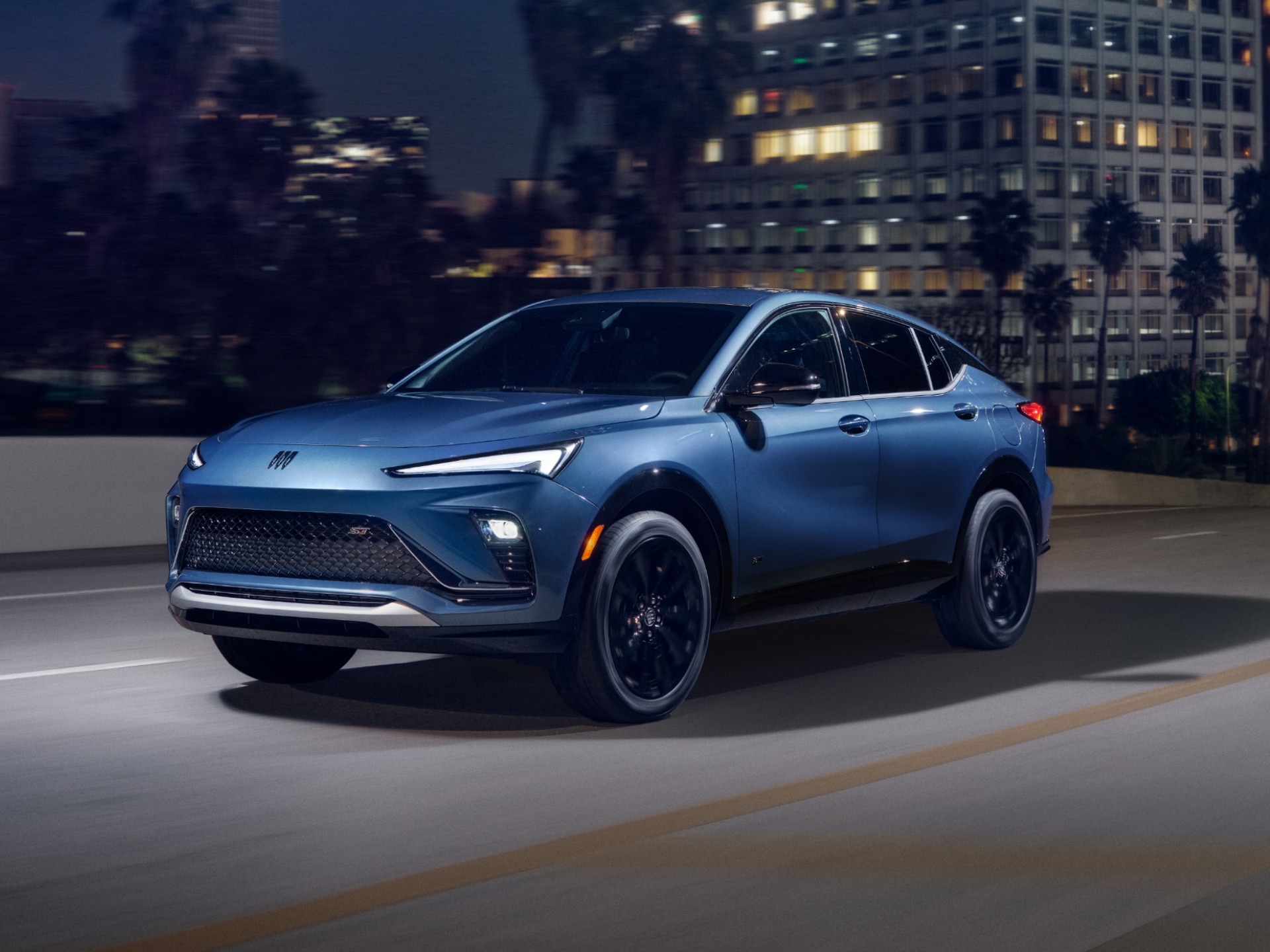 A Blue Buick Envista Compact SUV Driving on a City Street at Night with Its Headlights On