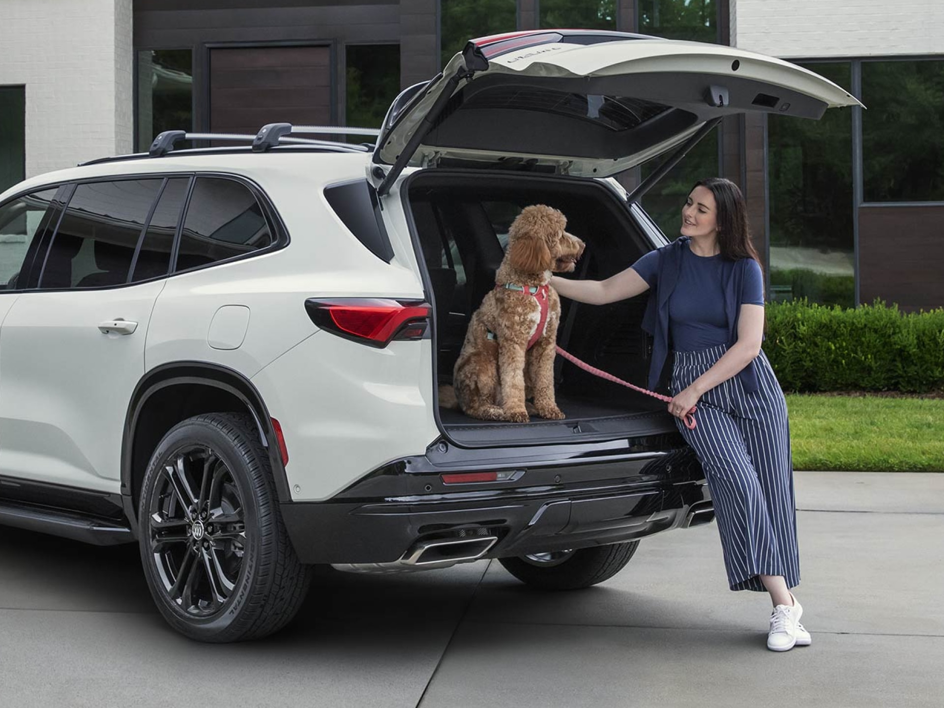 A Woman Sitting Beside the Open Liftgate of a White Buick Enclave SUV Petting a Brown Poodle Wearing a Red Harness