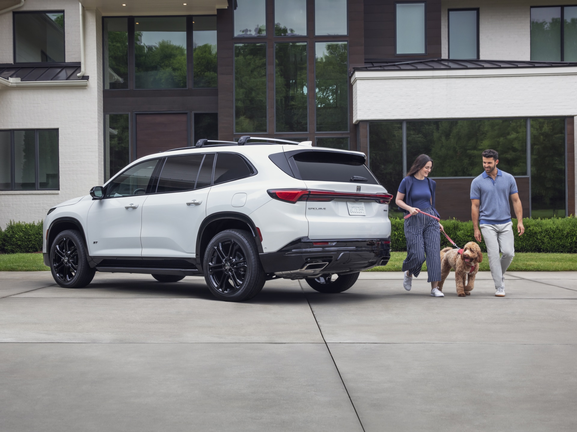A White Buick SUV with Black Wheels is Parked on a Driveway with a Couple and Their Dog Walking Away From It