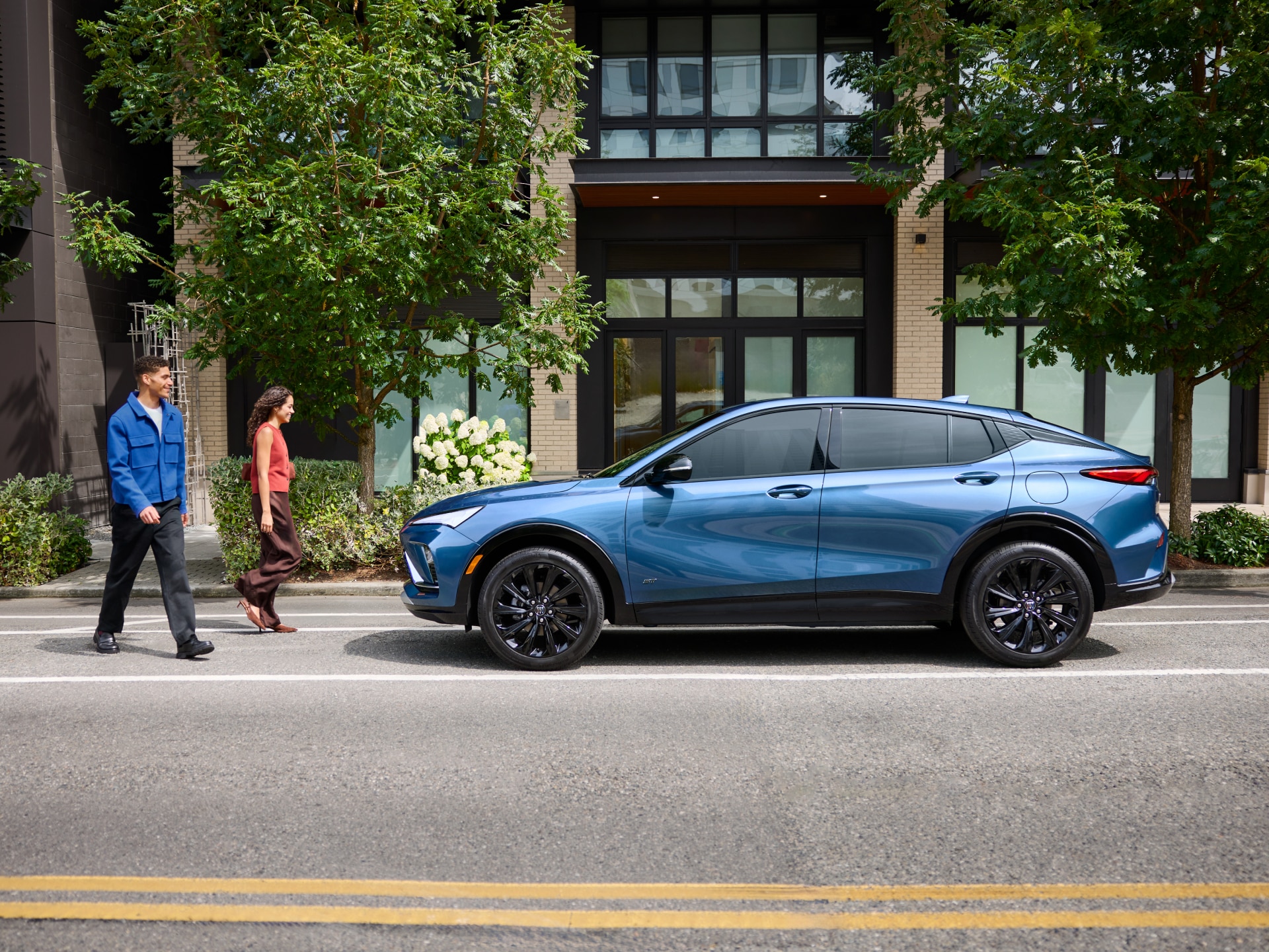 A Metallic Blue Buick Envista Parked on a City Street with a Couple Walking in Front of it and Green Foliage in the Background