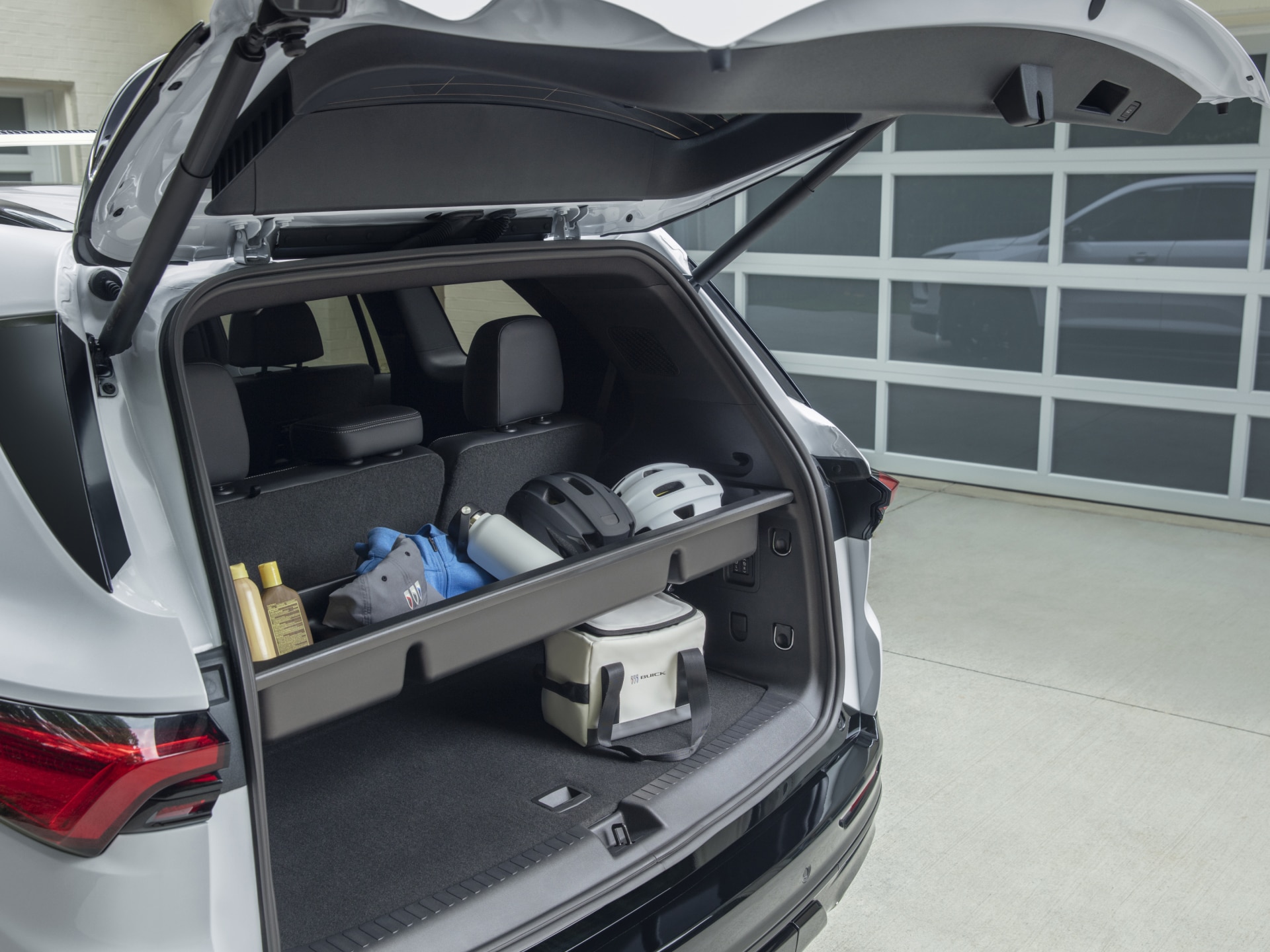 The Open Rear Cargo Area of a White Buick SUV with a Shelf Holding Helmets, Sunscreen, and a Buick Cooler Against a White Garage Door