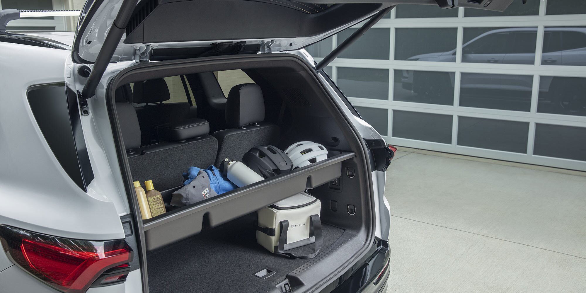 The Open Rear Cargo Area of a White Buick SUV with a Shelf Holding Helmets Sunscreen and a Buick Cooler Against a White Garage Door