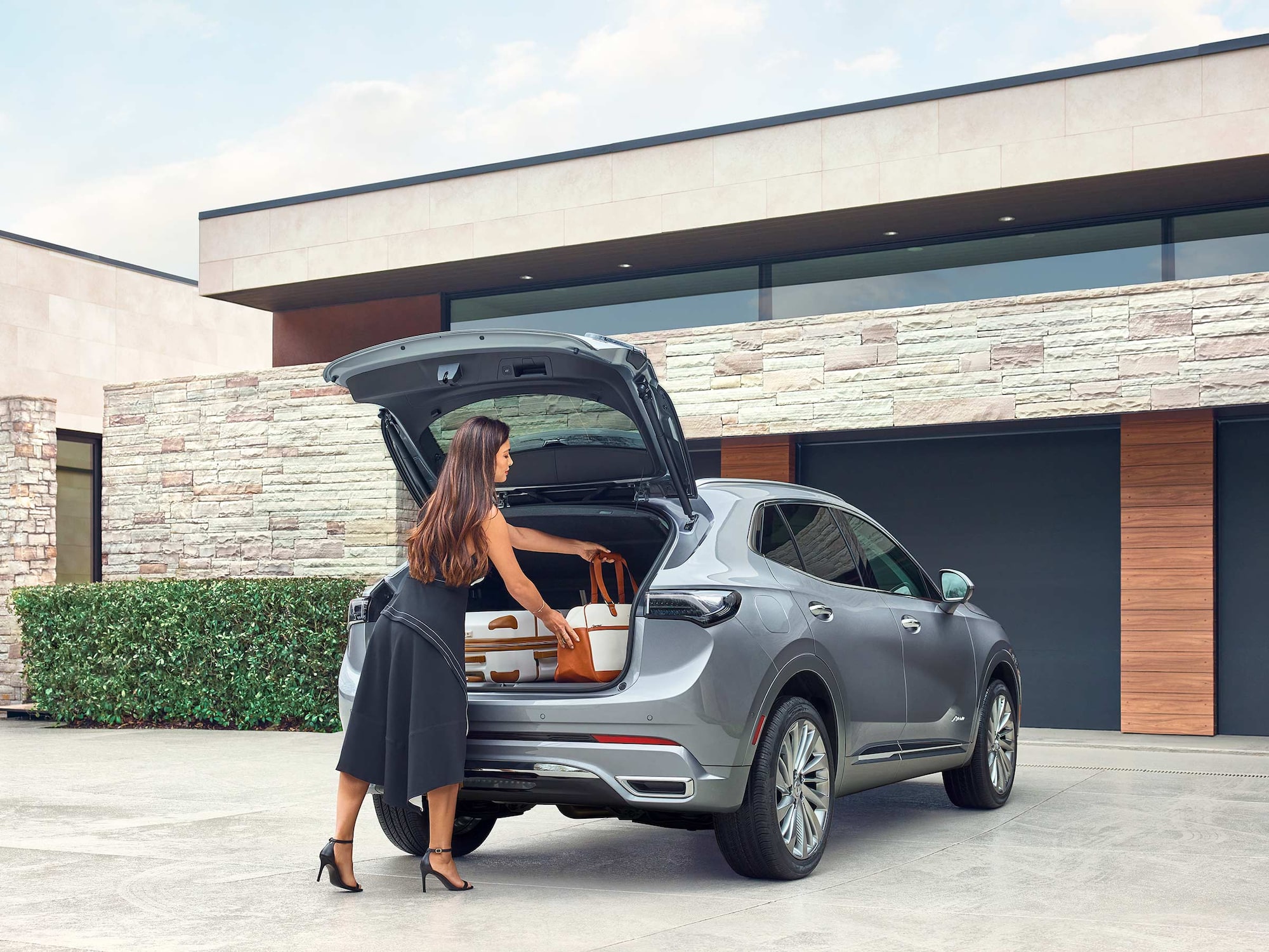 A Woman Loads Two Suitcases and a Handbag into the Open Trunk of a Gray Buick SUV Parked in a Driveway by a Stone House