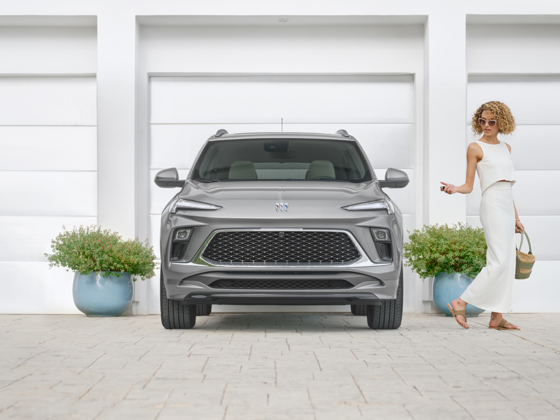 A Gray Buick SUV Parked in Front of White Garage Doors with a Woman in White Walking Away Holding a Key and a Bag